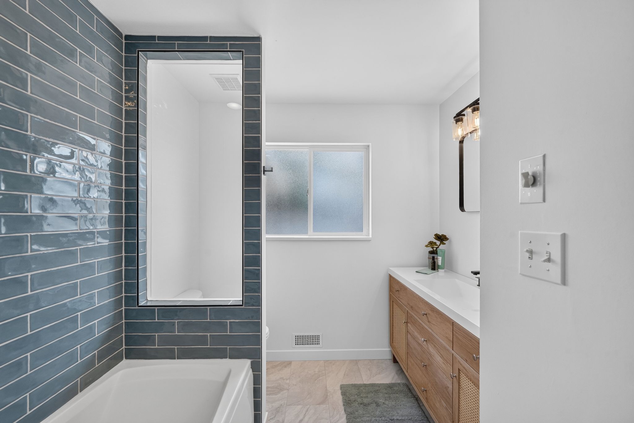 Bathroom featuring a deep soaking tub with blue subway tile surround, a built-in shower, a frosted window, a light wood-finish vanity with an integrated sink, and an elegant vanity light fixture