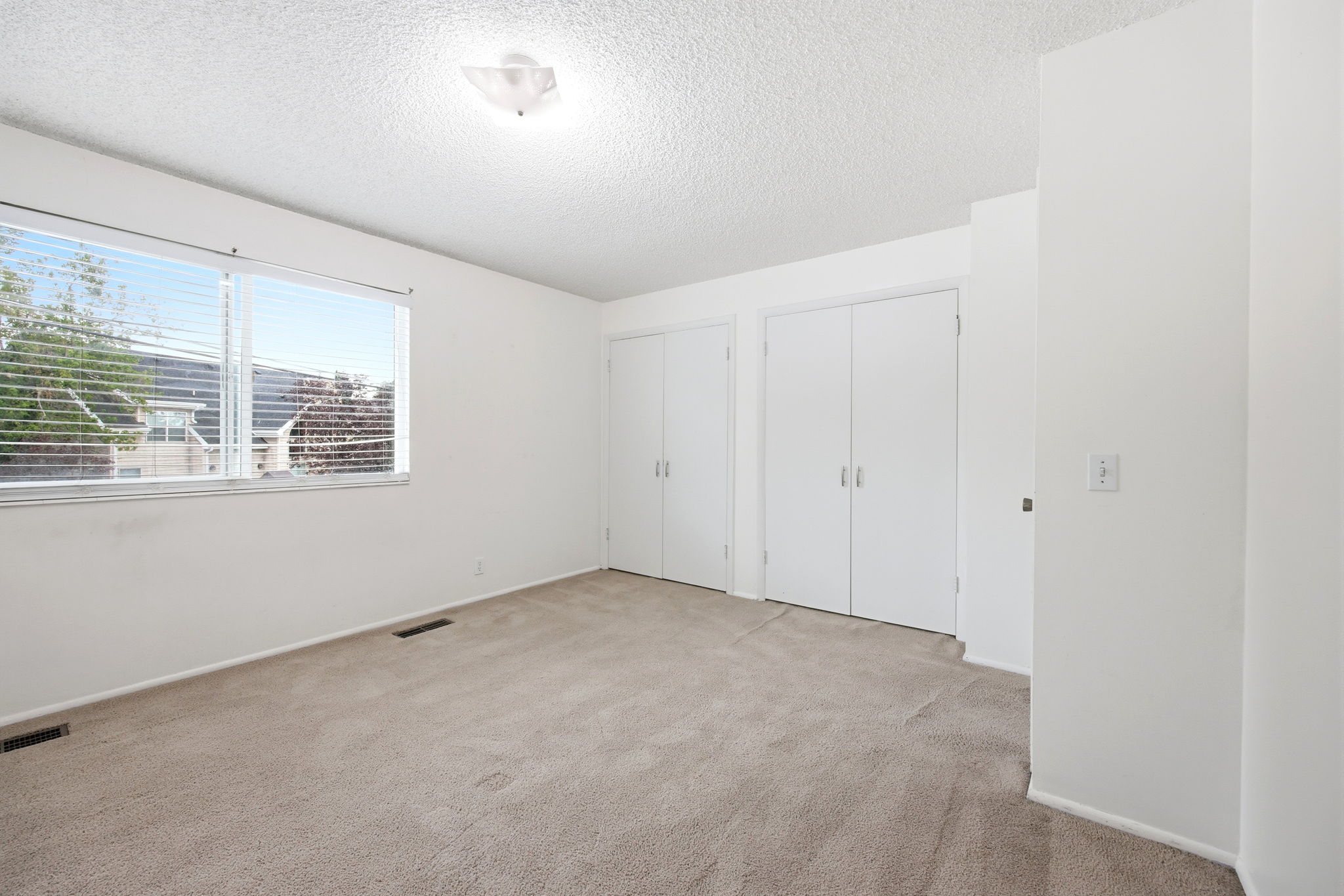 Neutral-toned carpeting spans this bright room, featuring a large window with horizontal blinds