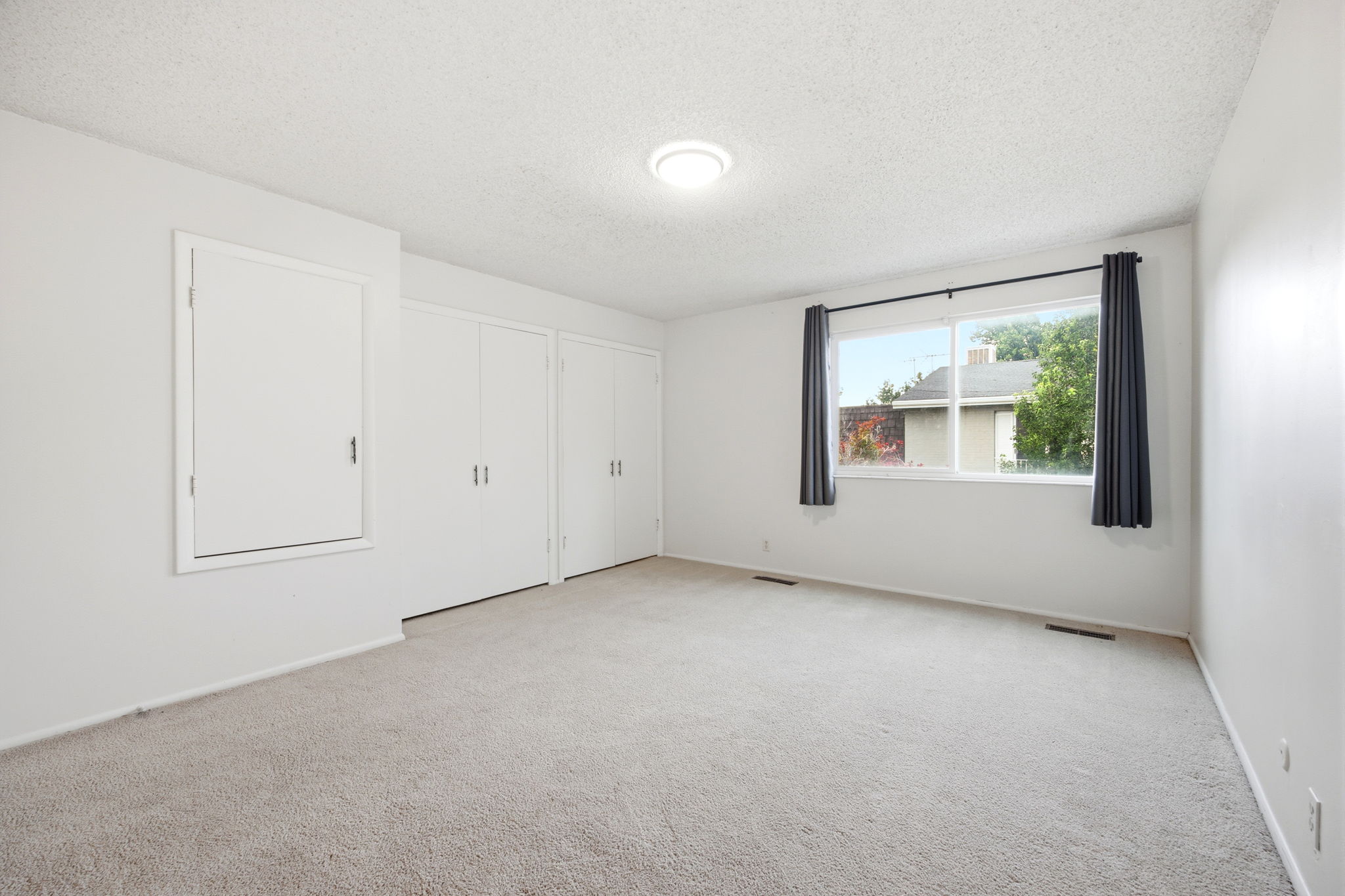 Bright interior featuring a large window, neutral carpeting, white walls, two sets of bi-fold closet doors, and a new ceiling-mounted light fixture