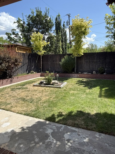 Fenced backyard featuring mature trees, a central planting bed, brick garden edging, and a concrete patio space