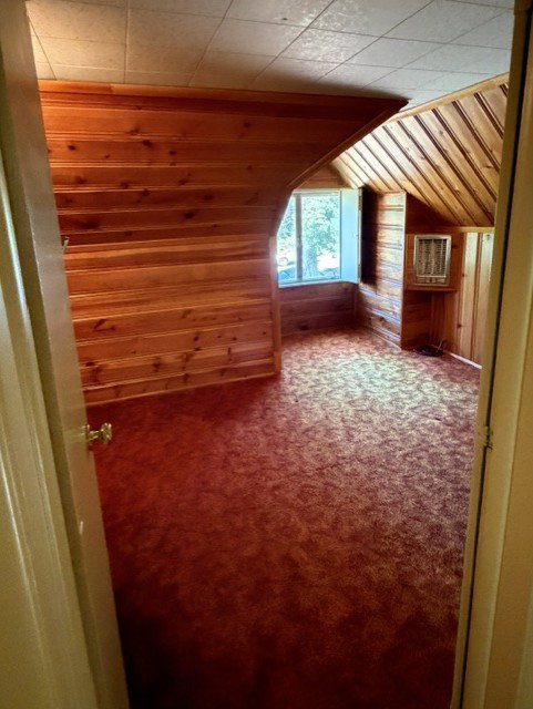 Bedroom with wood paneling on walls and ceiling, wall-mounted air conditioning unit, window, textured ceiling, and patterned red carpeting