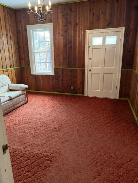 Room featuring extensive wood paneling, a white paneled door with upper glass inserts, a single white-framed window, and a five-light chandelier  Can be used for mail level bedroom
