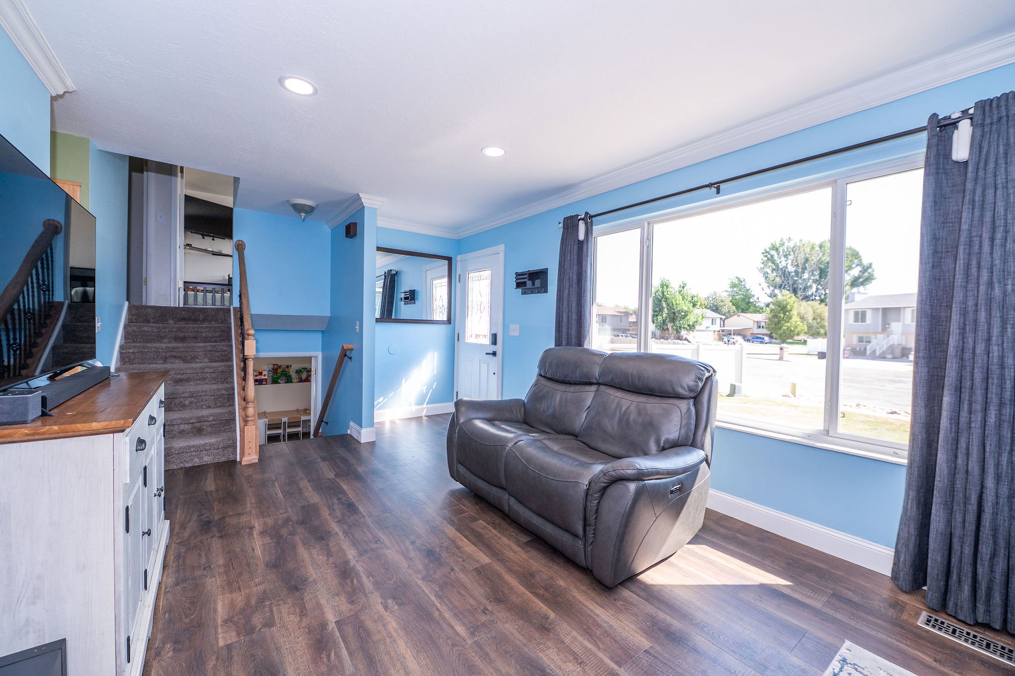 Spacious living area featuring large picture windows, wood-finish flooring, a white entry door with glass insert, and a staircase with wood newel posts and handrails