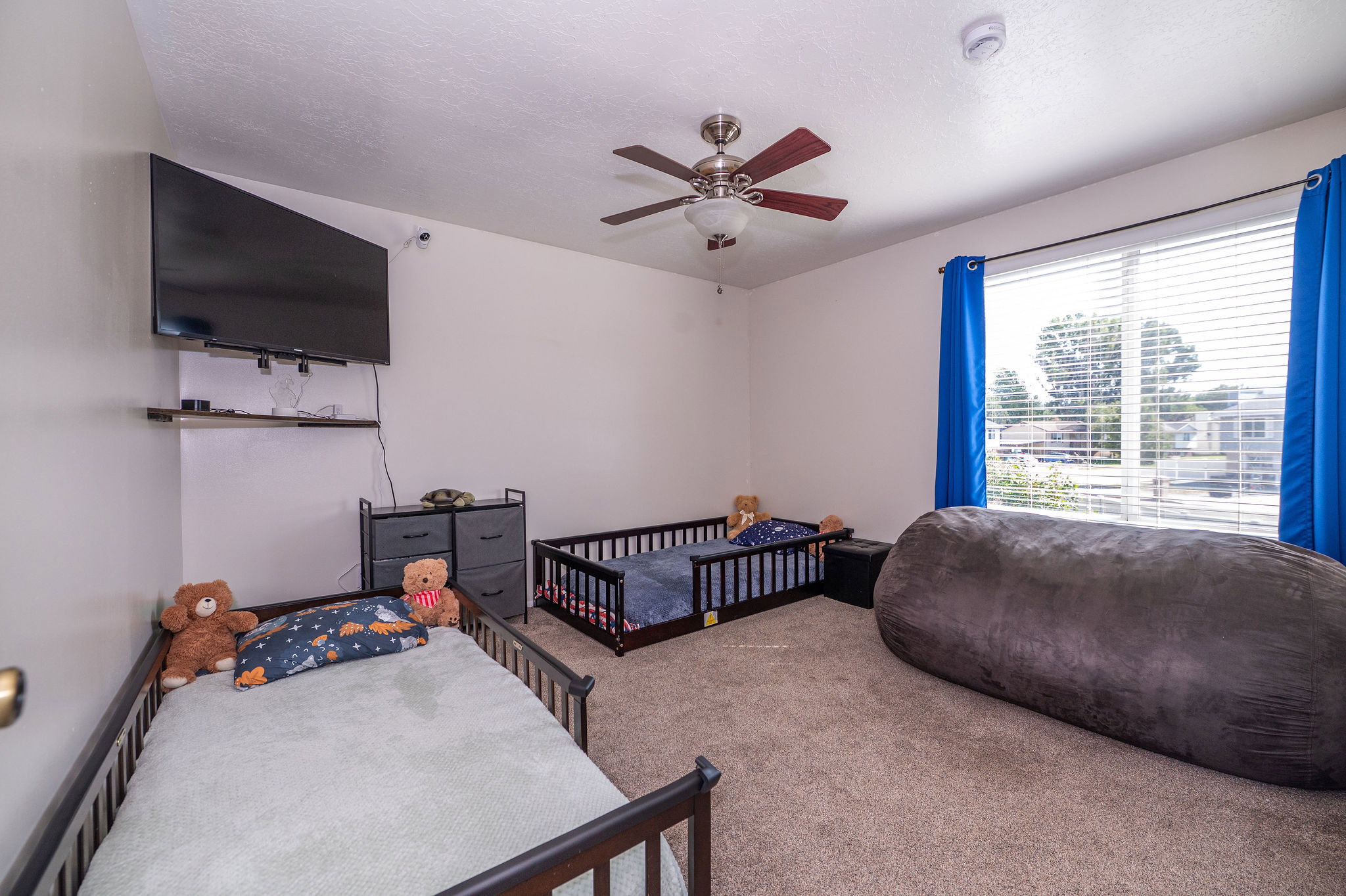 Carpeted room featuring a ceiling fan, a wall-mounted flat-screen television, a floating shelf, and a large window with horizontal blinds and blue drapes