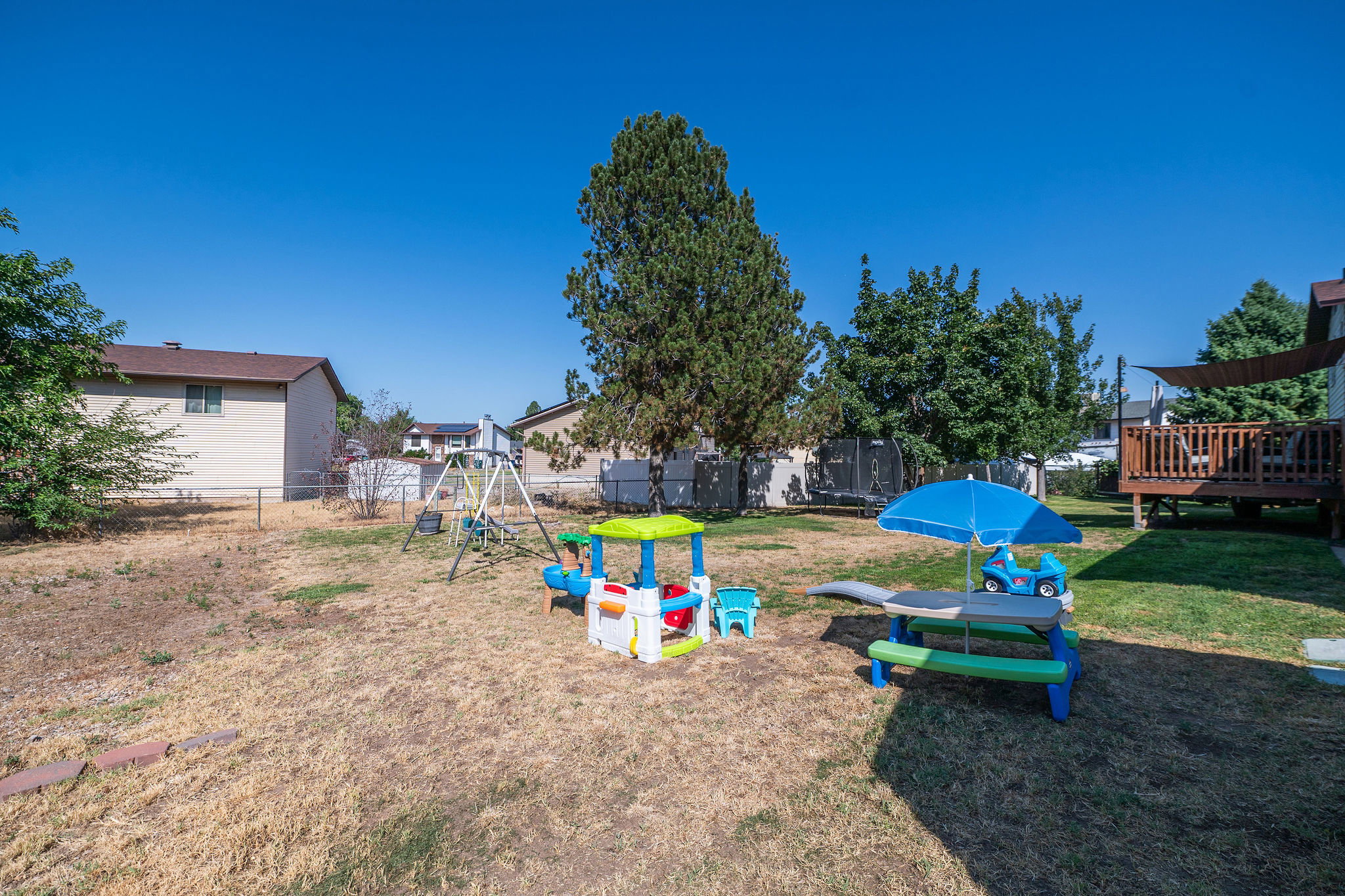 Expansive backyard featuring a raised wooden deck with a shade canopy, mature trees, and chain-link fencing