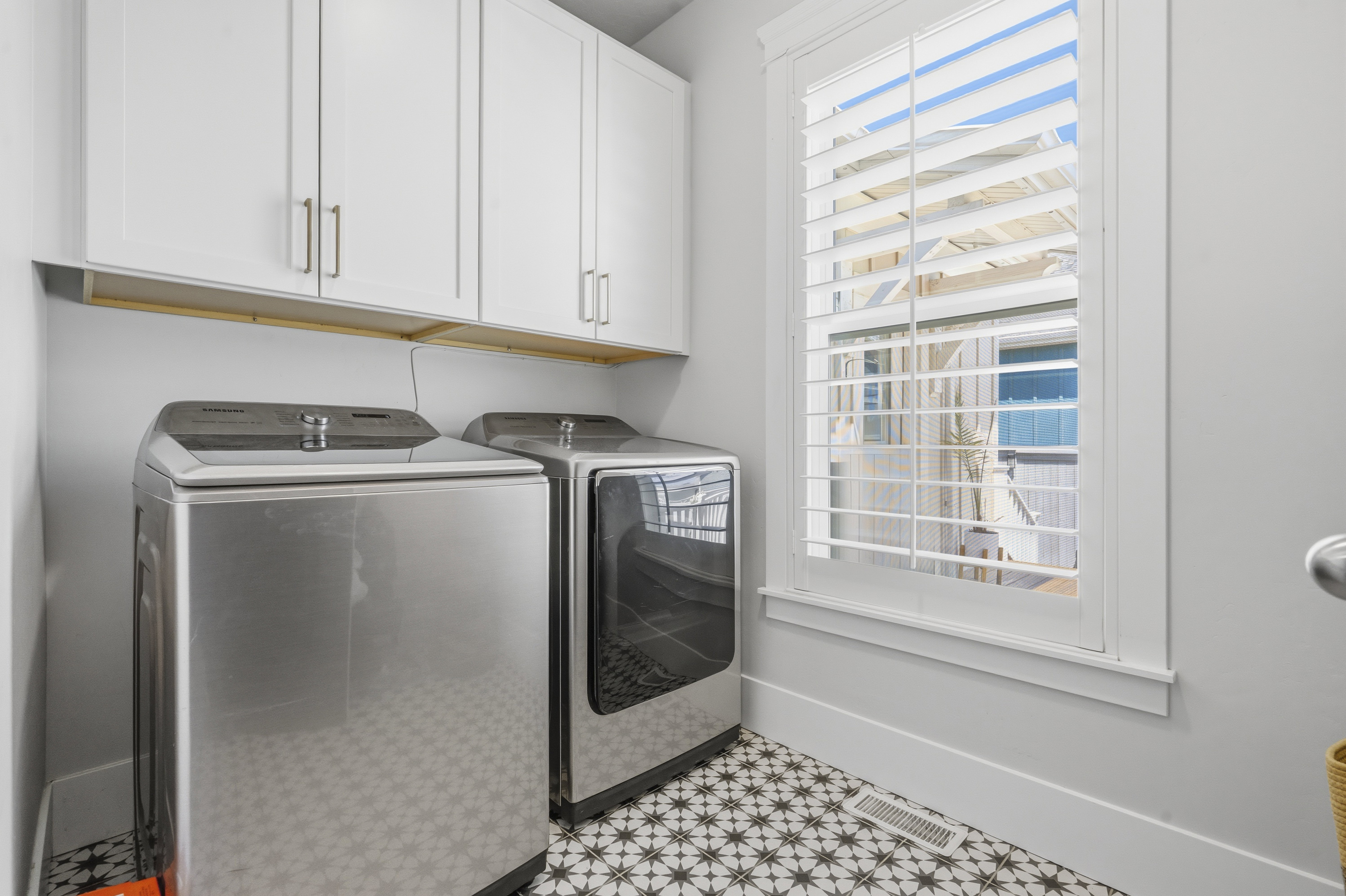 Laundry area featuring a window with white louvered shutters, patterned floor tiles, white shaker-style cabinetry with brushed nickel hardware, and an integrated wood shelf