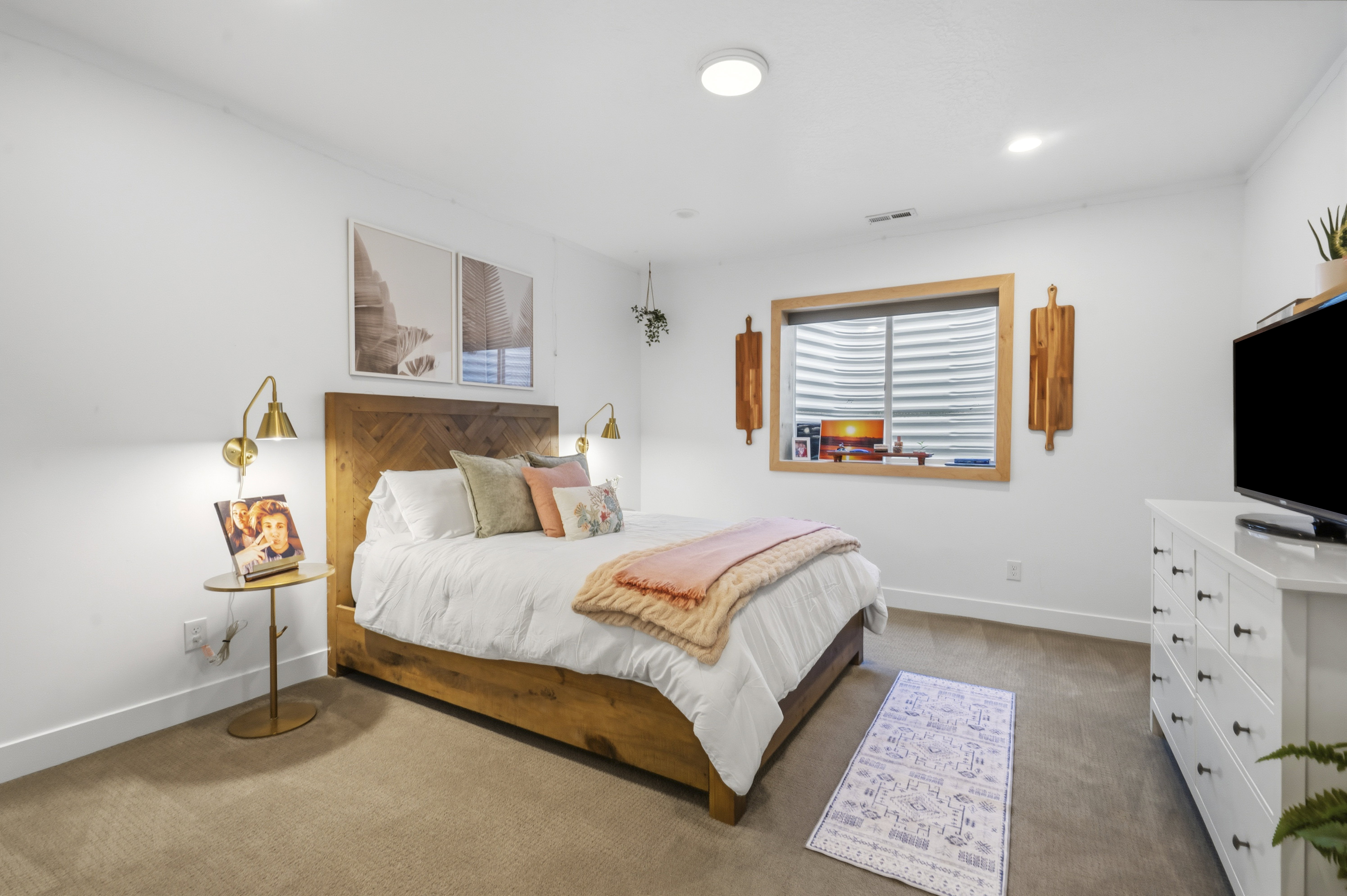 Carpeted room featuring white walls, a wood-finish bed frame with a herringbone headboard, two brass-finish wall sconces, and a window with wood trim