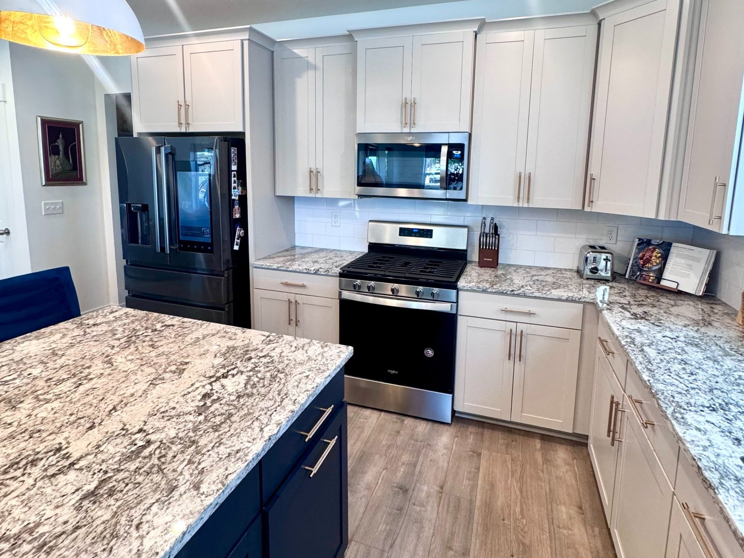 Contemporary kitchen featuring an expansive center island with dark cabinetry, light-toned perimeter cabinetry, stainless steel appliances, speckled countertops, and wood-finish flooring