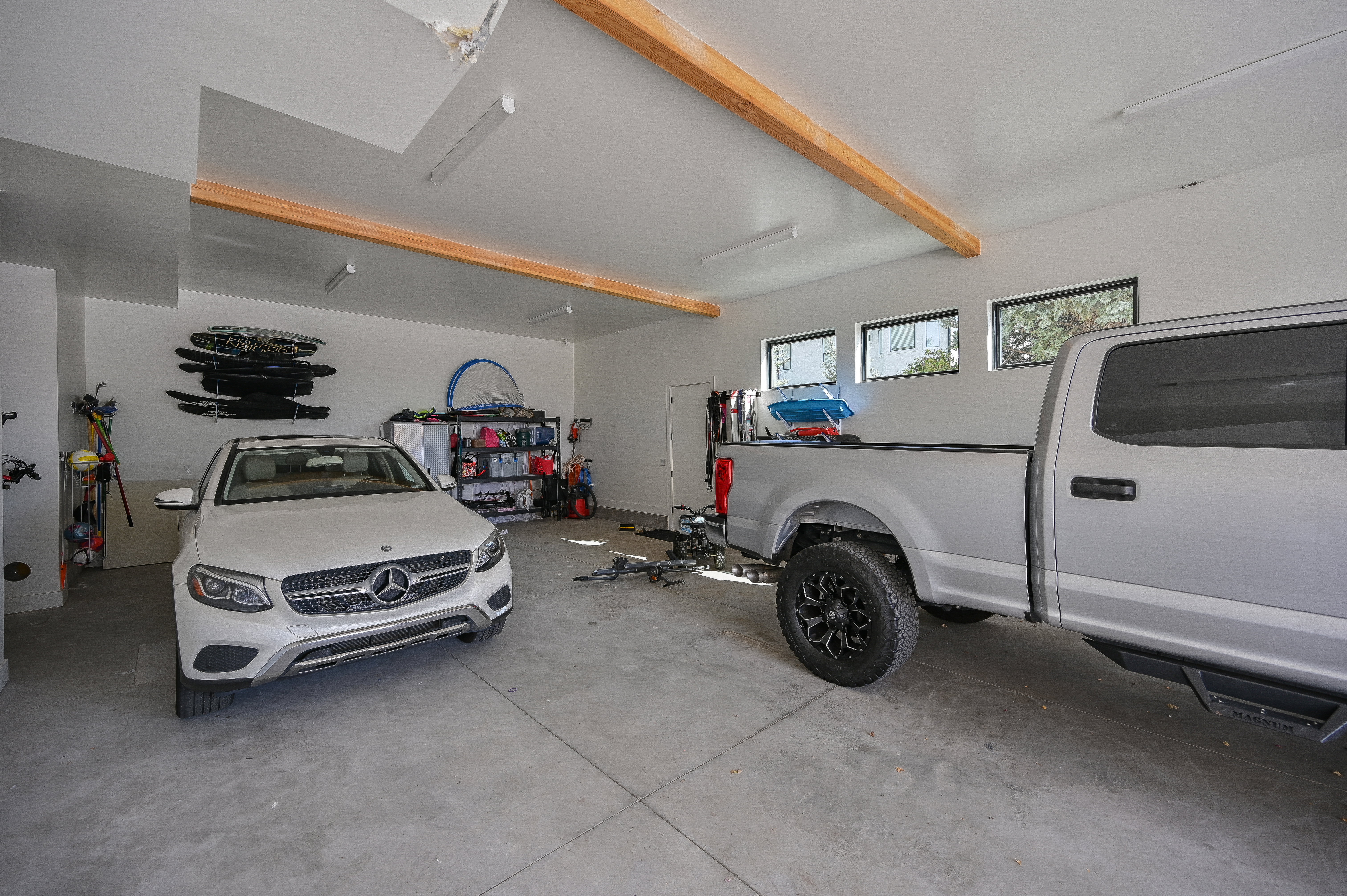 Spacious two-car garage featuring a concrete floor, white walls, exposed wood beams, and multiple windows providing natural light