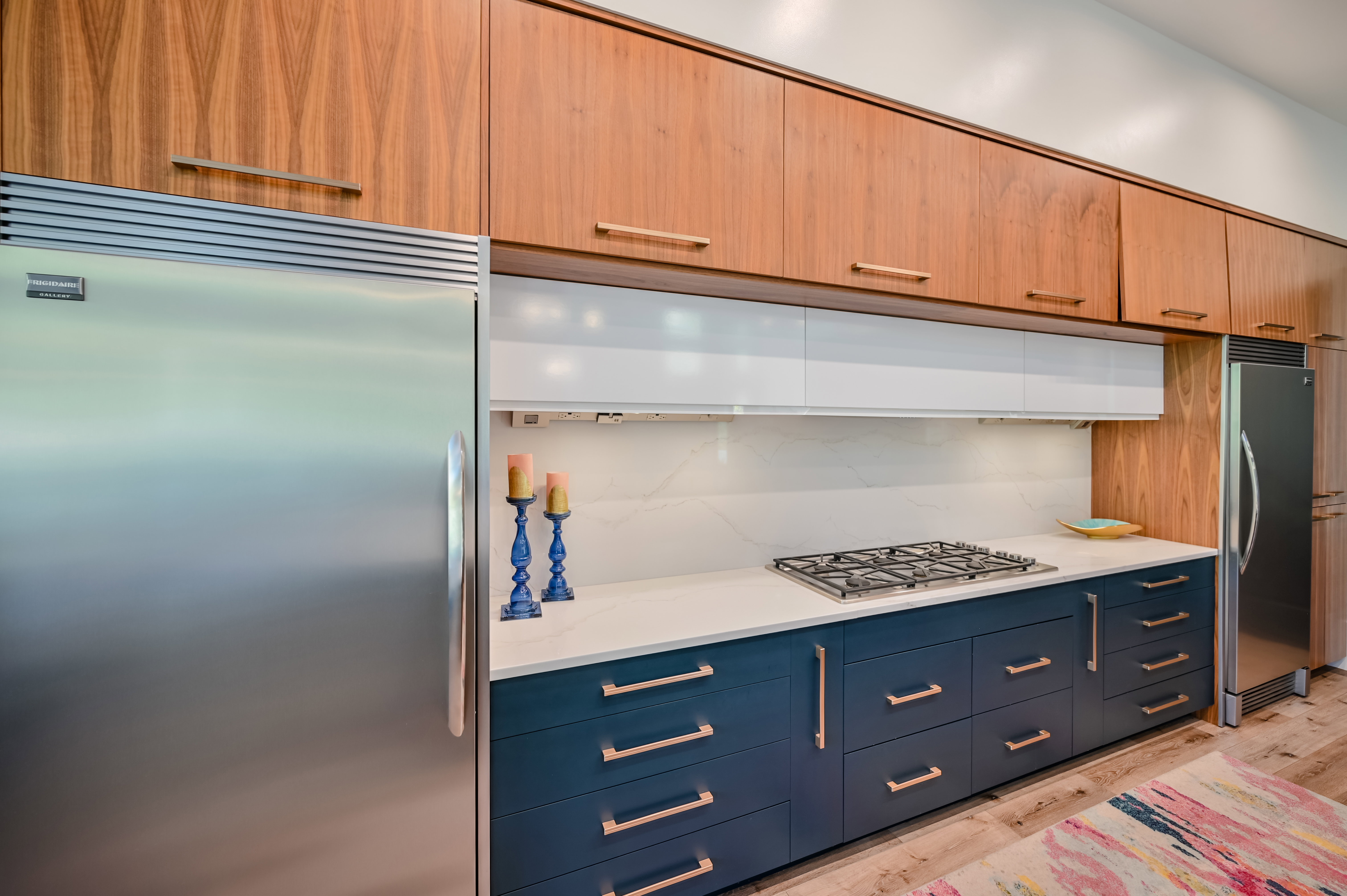 Contemporary kitchen featuring two-tone cabinetry with wood-finish upper cabinets and deep blue lower cabinets, stainless steel appliances, a built-in gas cooktop, and a light-toned countertop with a matching backsplash