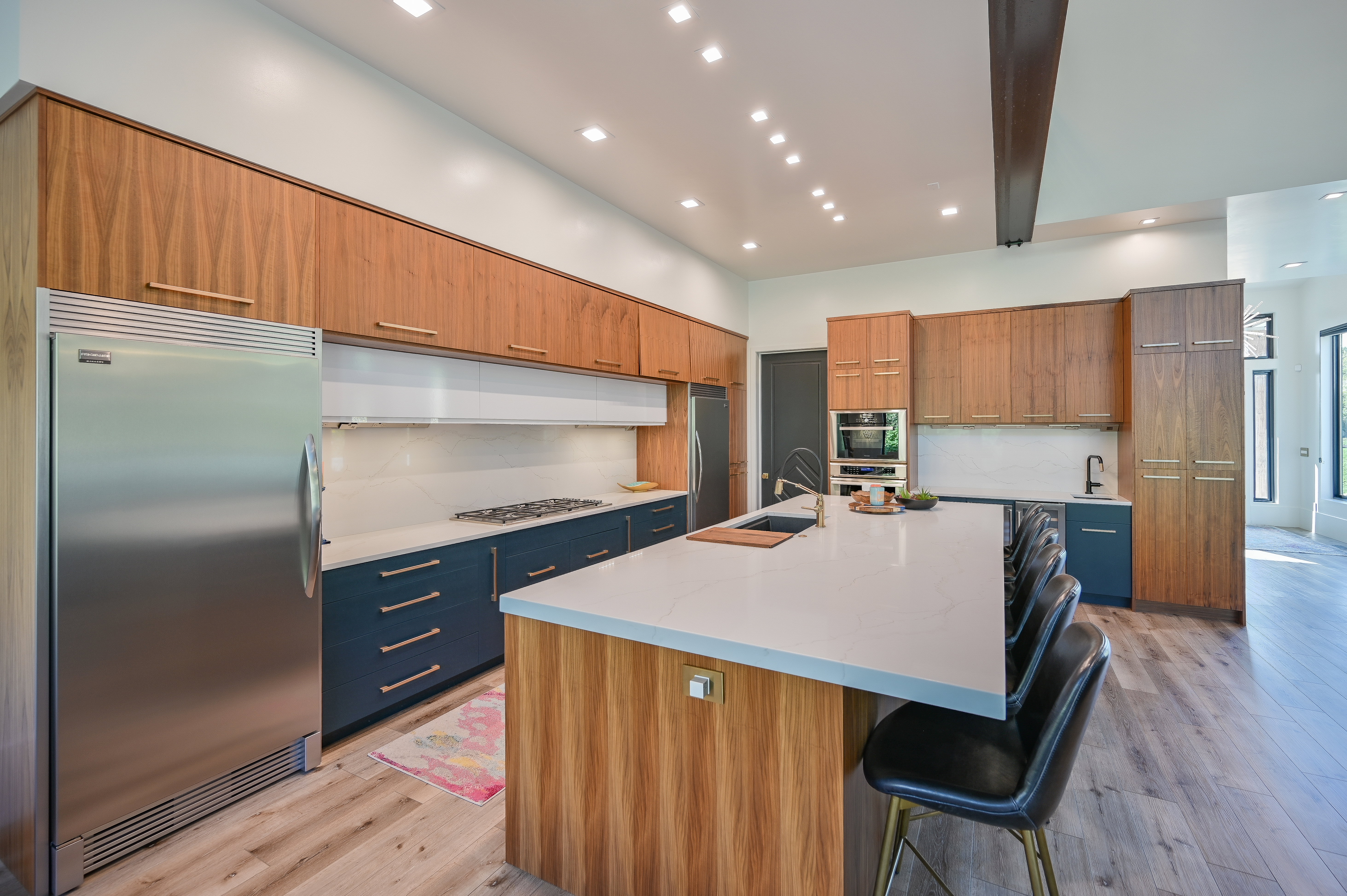 Modern kitchen featuring light wood-finish upper cabinetry, blue lower cabinetry, stainless steel appliances, a white countertop island, and recessed lighting