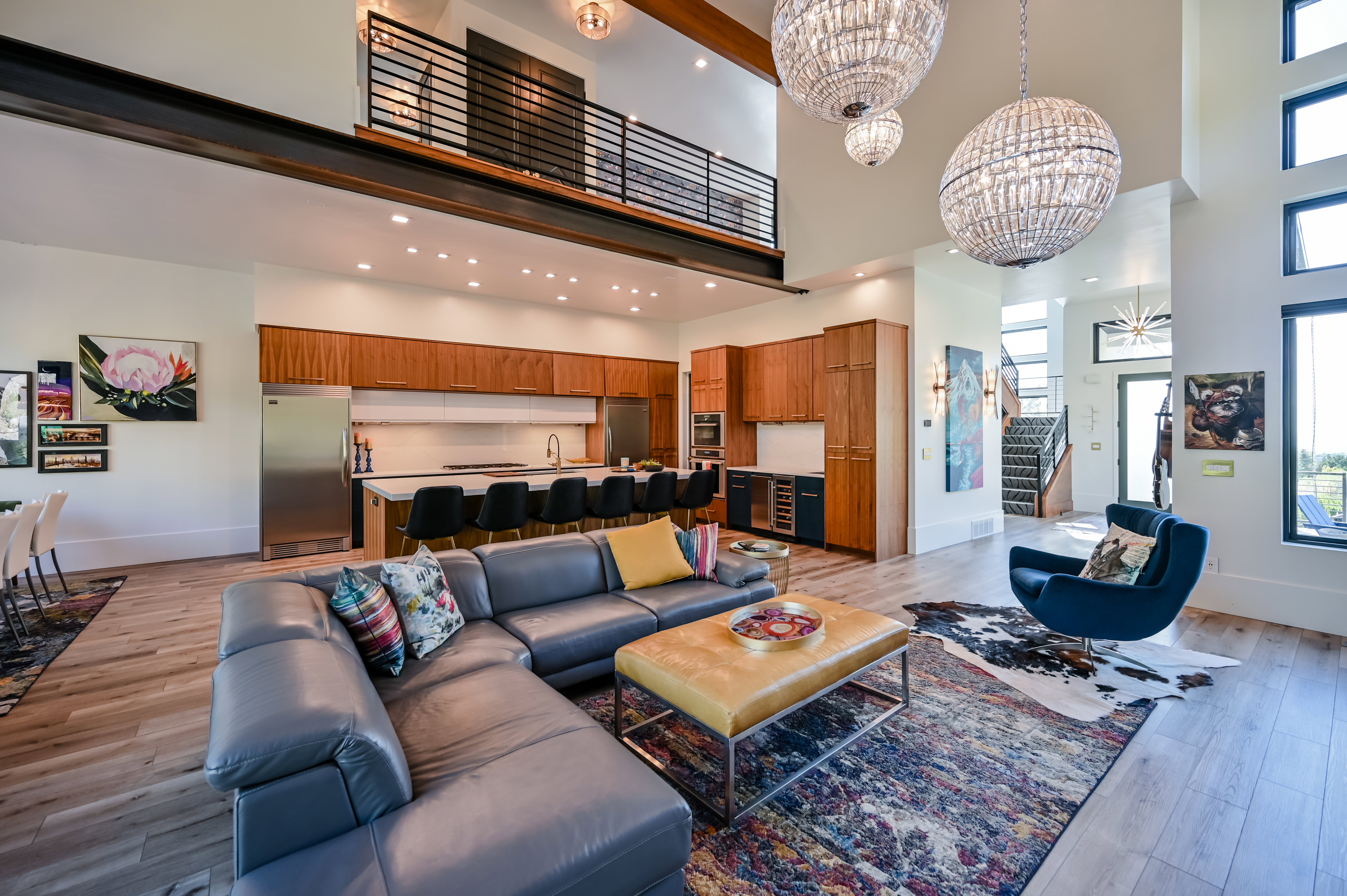 Spacious open-concept living area featuring a double-height ceiling, wood-finish flooring, a contemporary kitchen with a large island, and a prominent second-story balcony with industrial-style railings