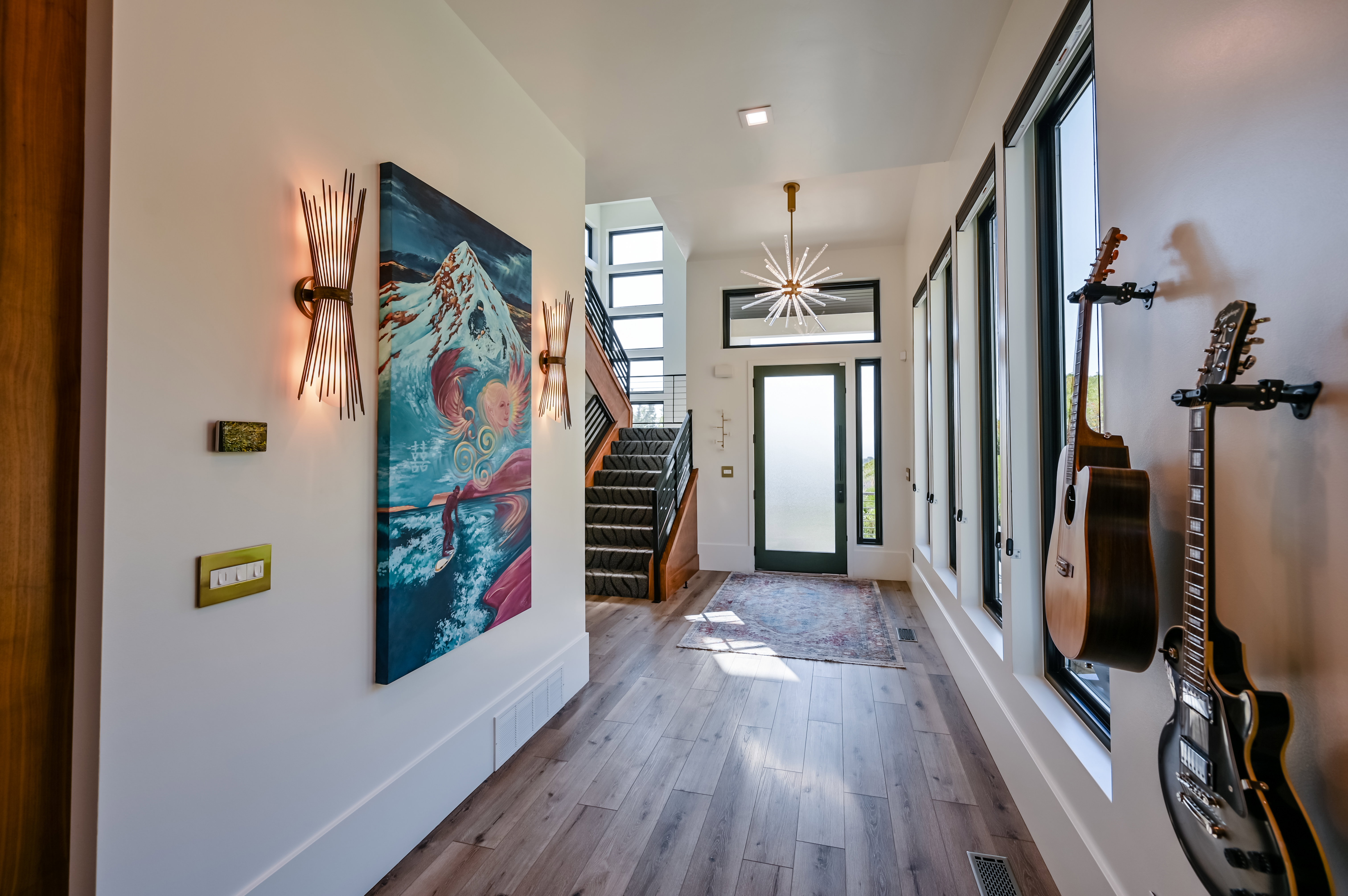 Open-concept entryway featuring wood-finish flooring, a contemporary chandelier, multiple vertical windows with dark trim, a prominent staircase with dark risers, and modern wall sconces