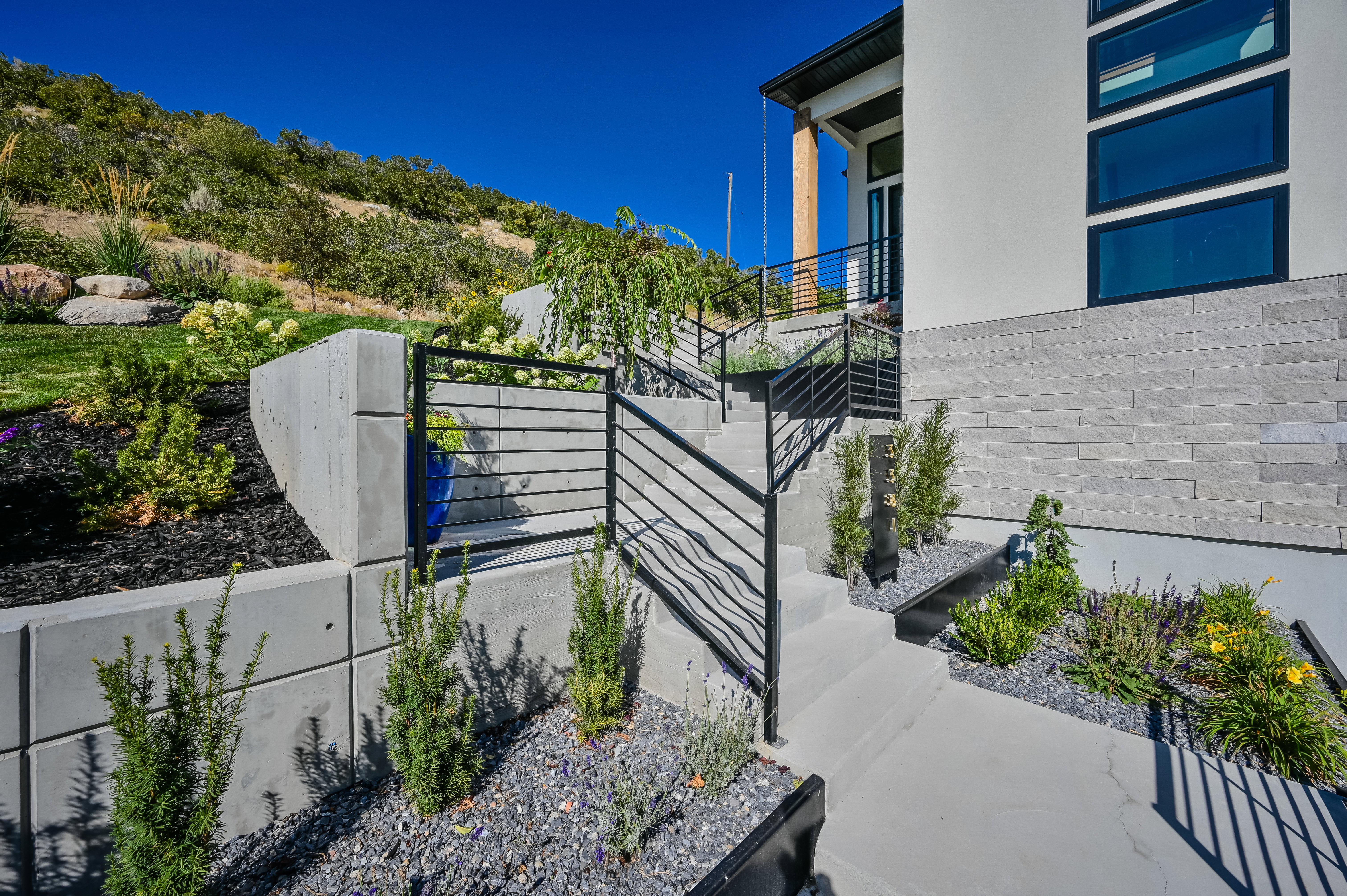 Modern exterior featuring stacked stone accents, multiple large windows, and a tiered concrete walkway with contemporary black railings