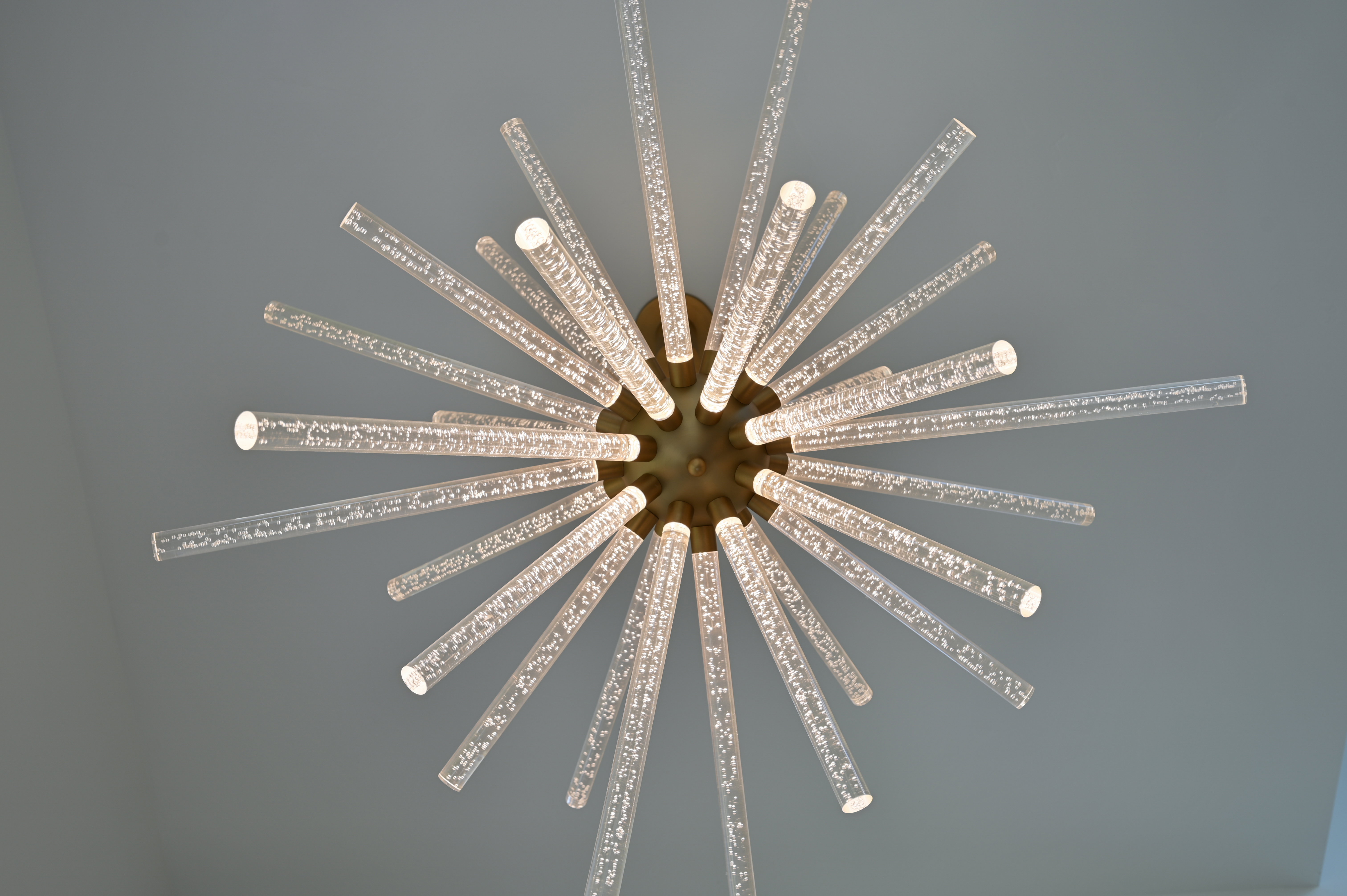 Contemporary chandelier featuring multiple radiating glass rods with internal bubble detailing, mounted on a central bronze-toned fixture against a smooth ceiling