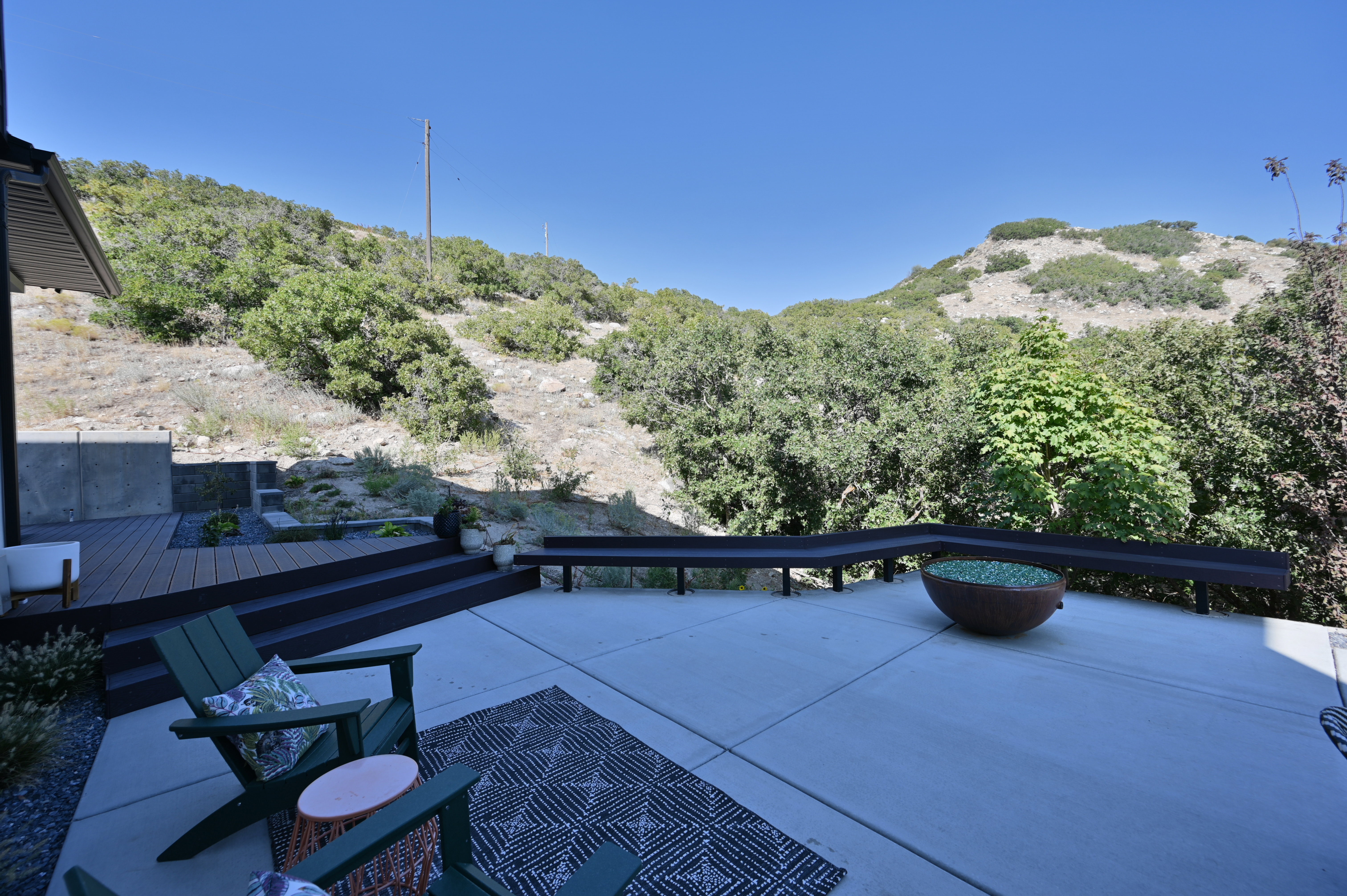 Expansive concrete patio with a tiered composite deck, built-in bench seating, a fire bowl feature, and surrounding mature foliage