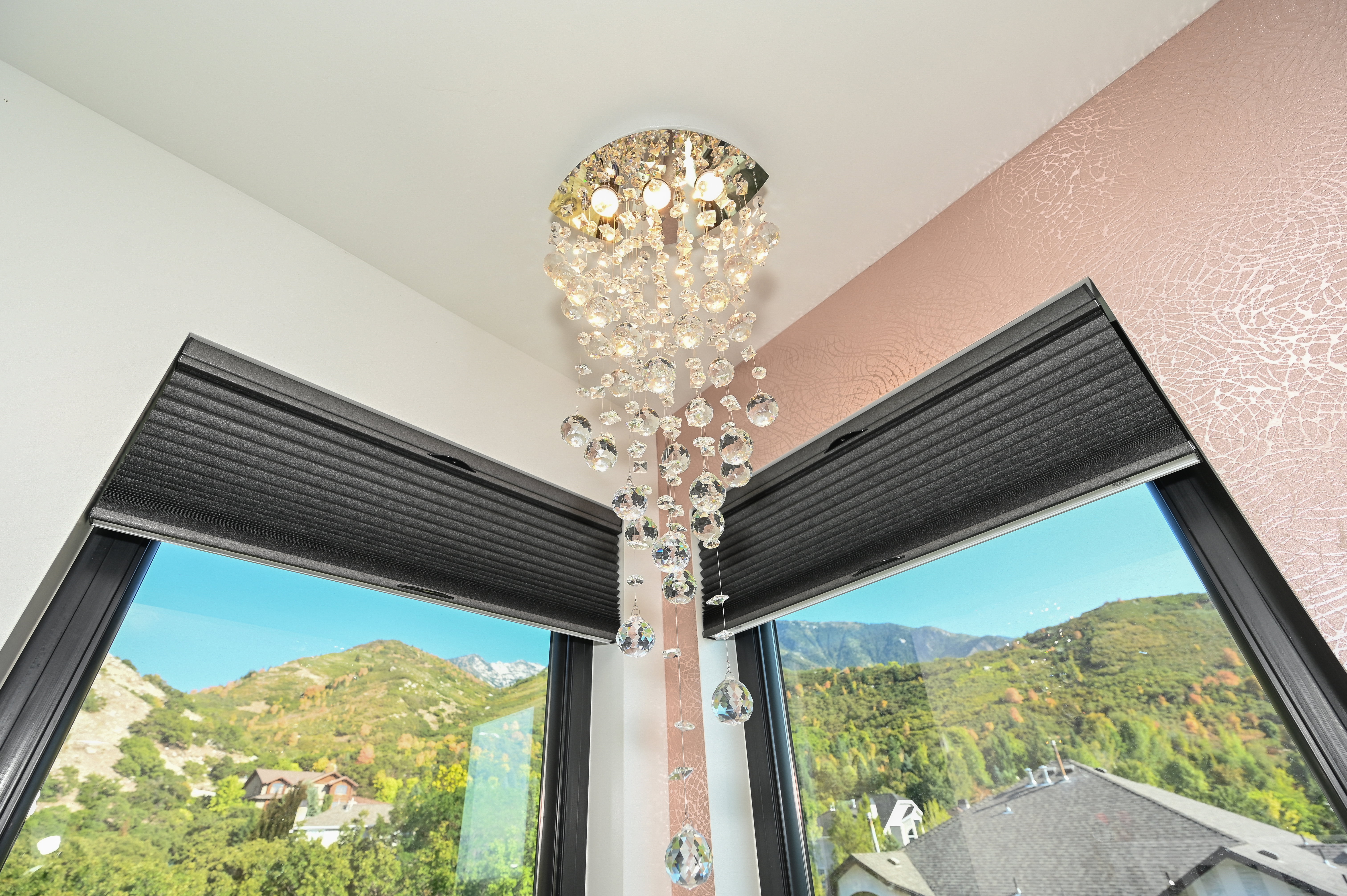 Ornate crystal chandelier illuminating an interior corner with textured rose-gold wallpaper and two large windows featuring black pleated shades