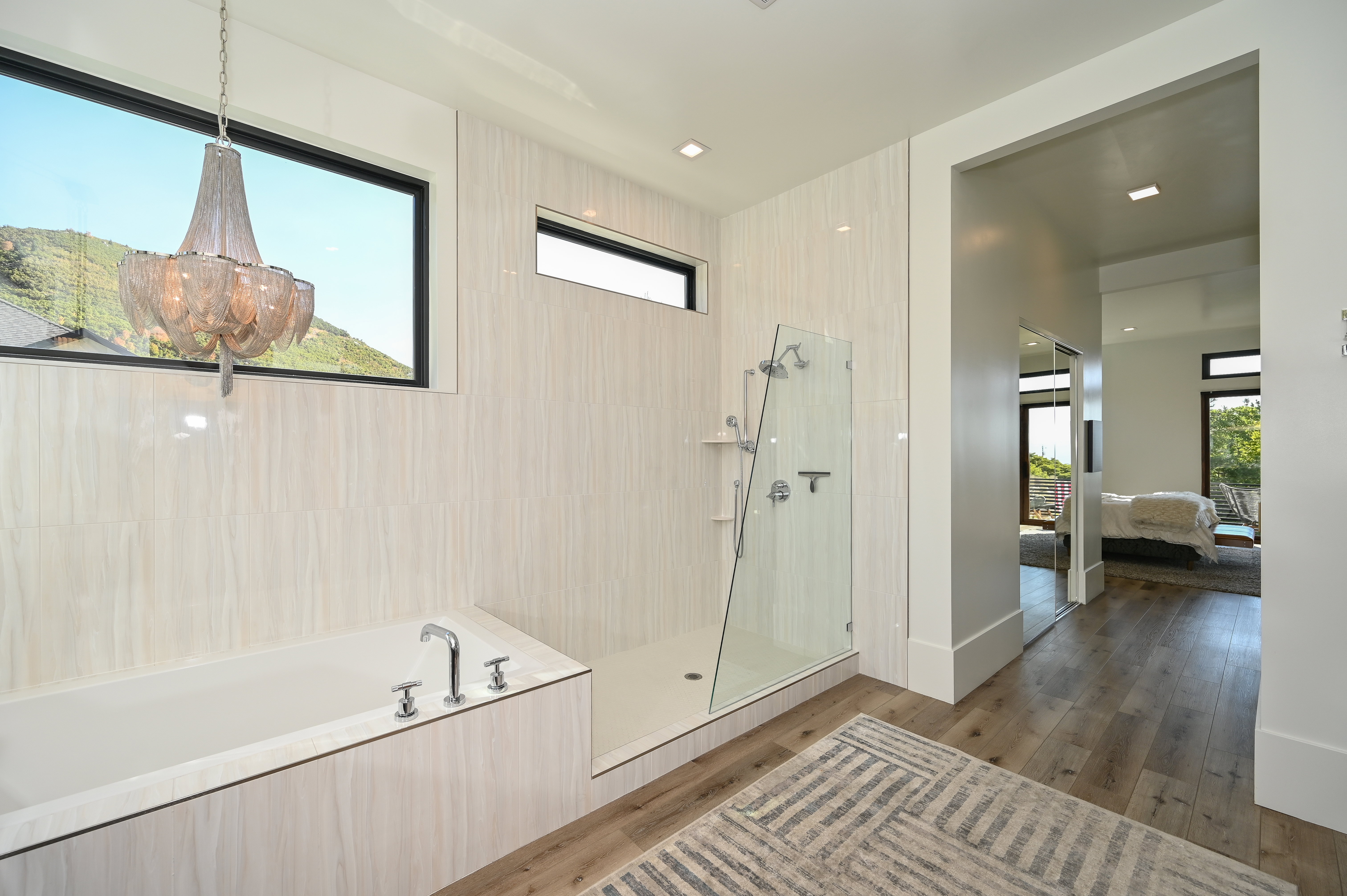 Contemporary bathroom featuring a built-in soaking tub, a frameless glass shower enclosure, wood-finish flooring, and a large window with a mountain backdrop