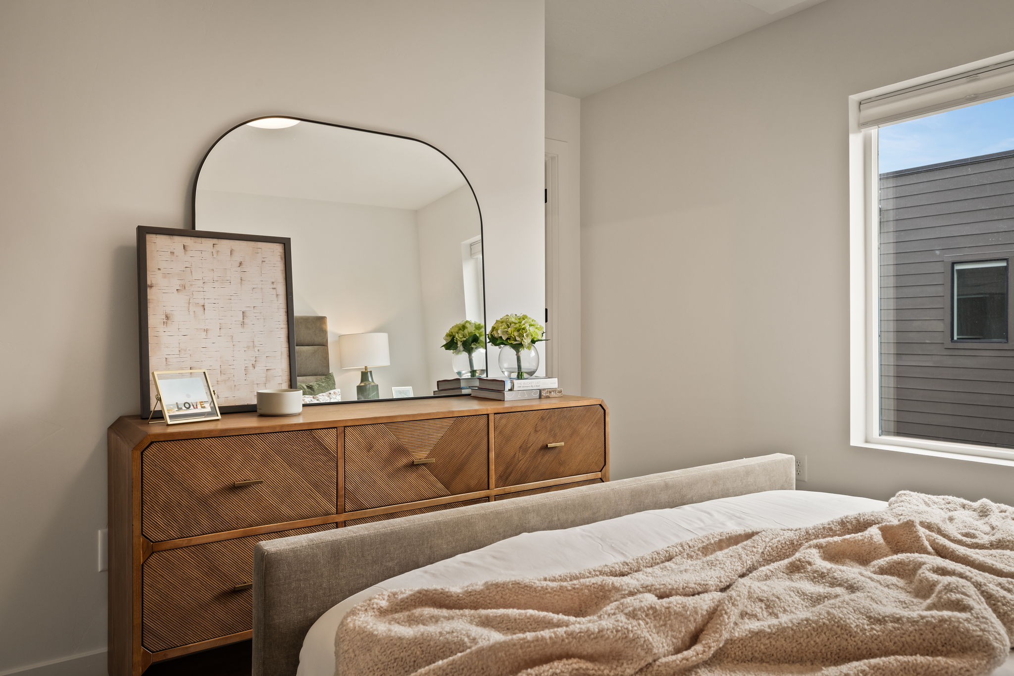 Bedroom featuring a wood-finish dresser with chevron-patterned drawers, a sleek arched mirror, and a large window with white trim