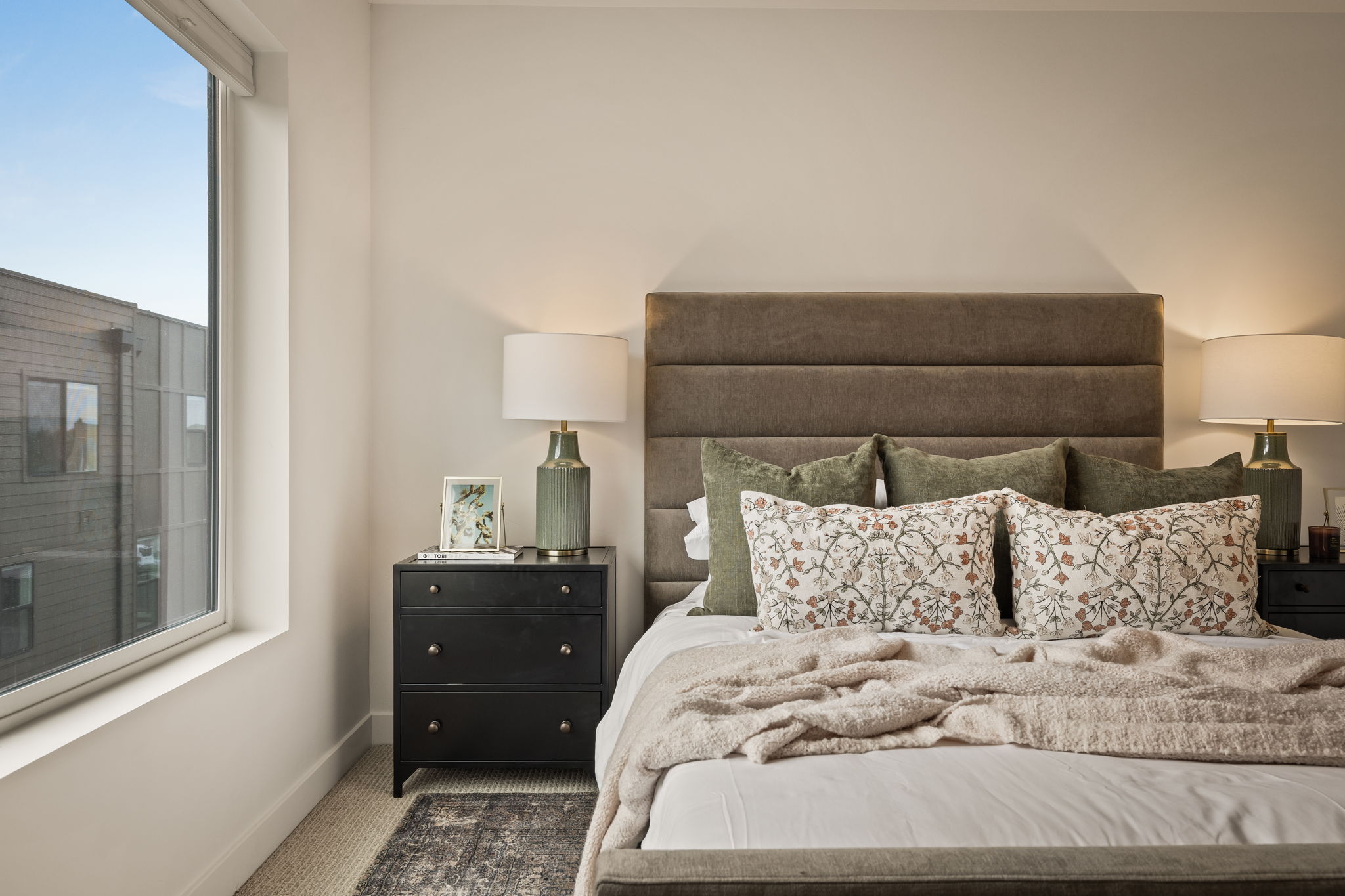 Bedroom featuring a large window, a paneled upholstered headboard, a dark wood nightstand with drawer pulls, and wall-to-wall carpeting