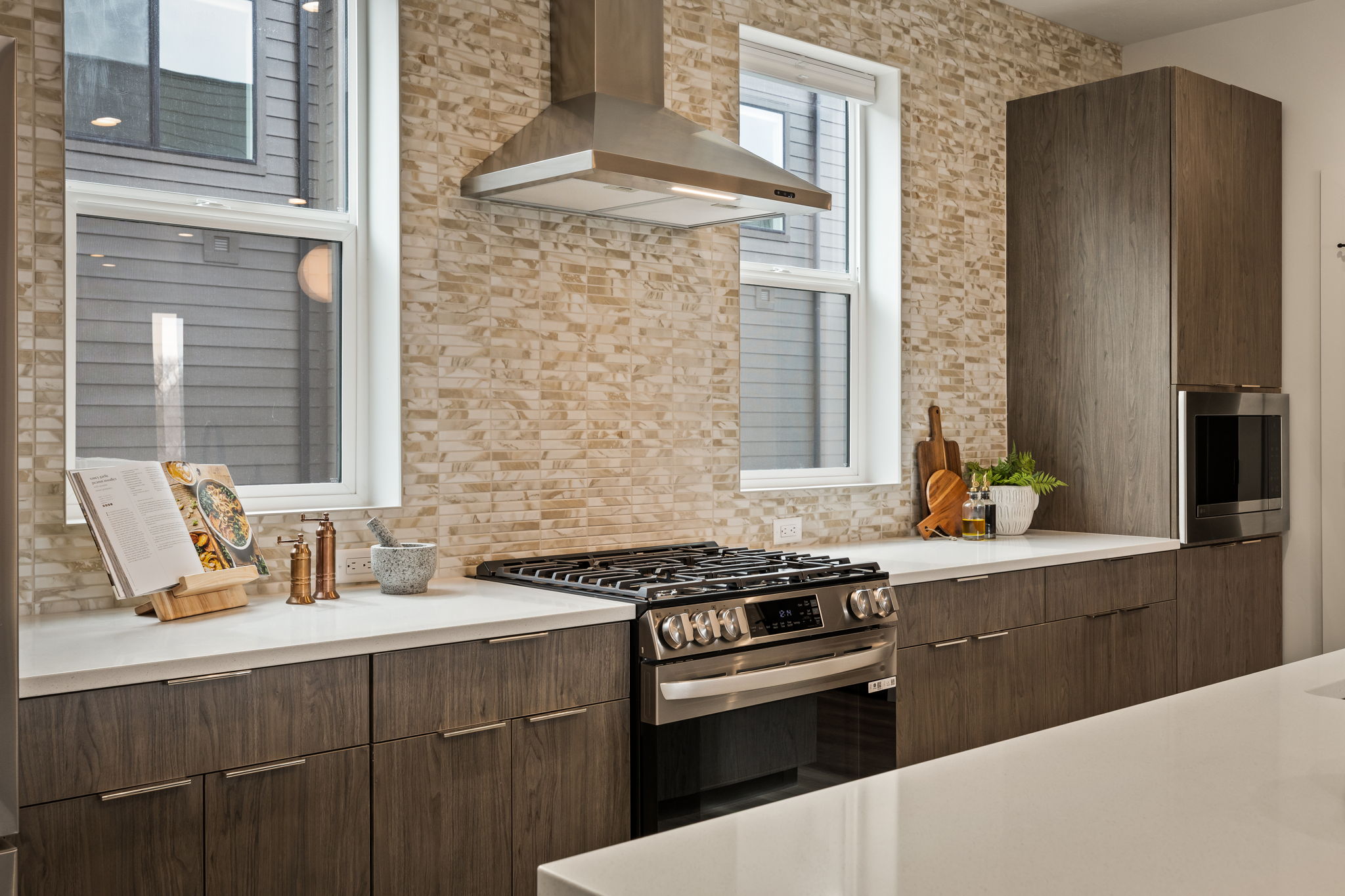 Modern kitchen featuring a stainless steel range with a coordinating hood, stone-finish backsplash, wood-grain cabinetry, light-toned countertops, and dual windows