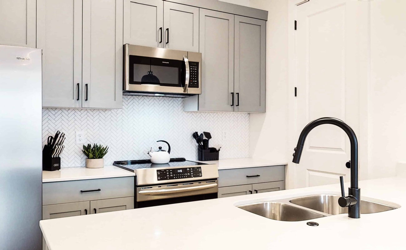 Modern kitchen featuring light gray cabinetry with black hardware, stainless steel appliances, white herringbone tile backsplash, and light-toned countertops