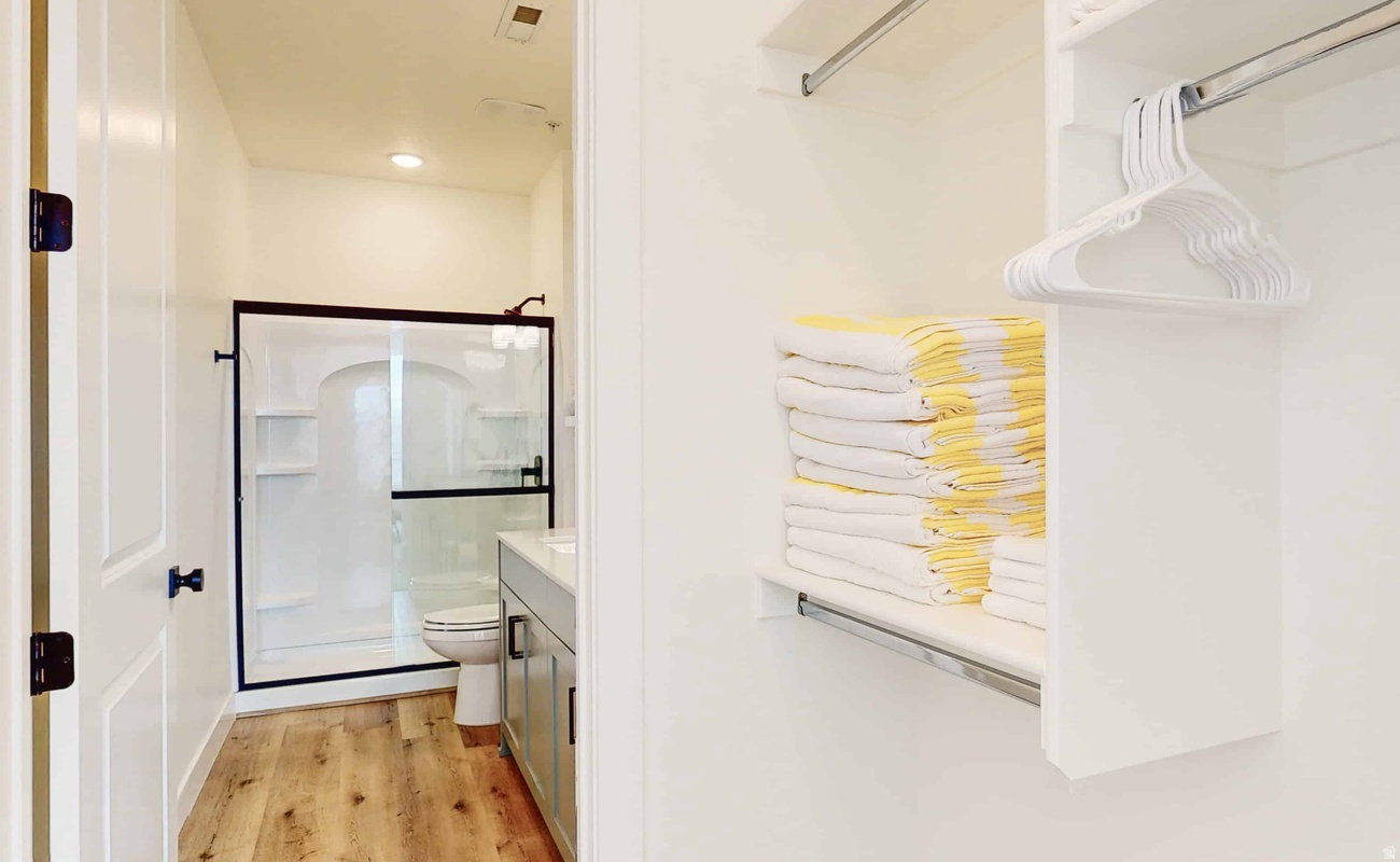 Built-in closet shelving and hanging rods with an adjacent bathroom featuring wood-finish flooring, a white vanity with a light-colored countertop, and a shower with a clear glass enclosure and dark-framed accents