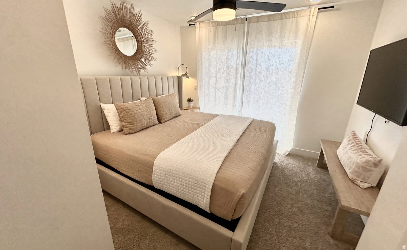 Plush carpeted flooring, a paneled upholstered headboard, and a ceiling fan with integrated lighting