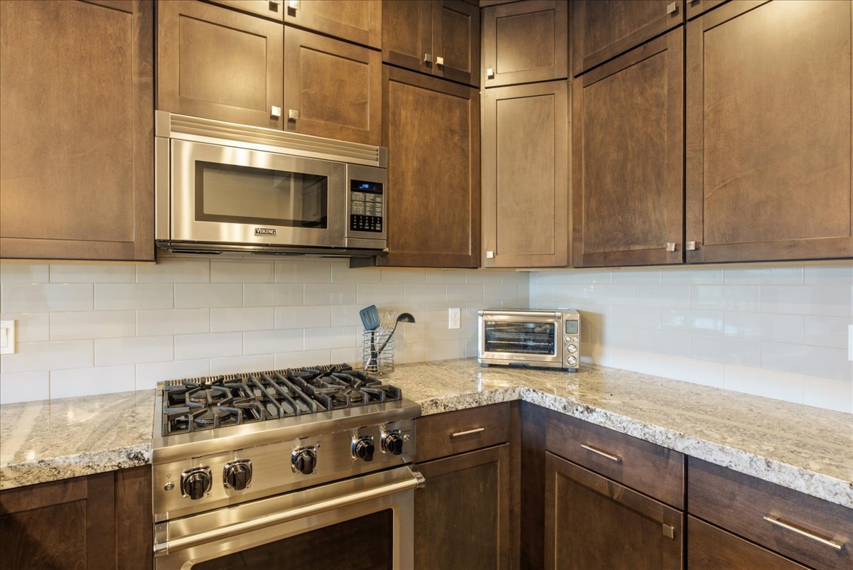 Kitchen featuring stainless steel range with overhead microwave, natural stone countertops, full-height tile backsplash, and extensive wood cabinetry