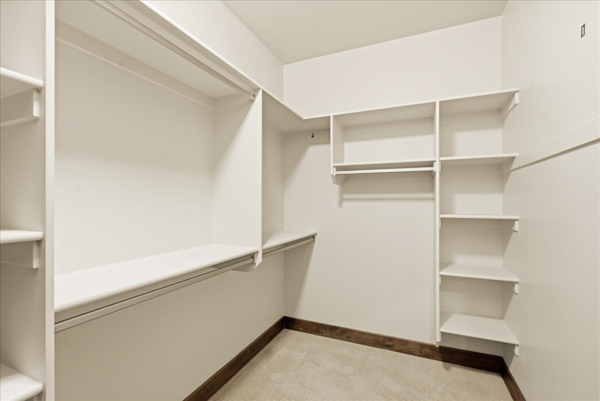 Walk-in closet featuring extensive built-in shelving, multiple hanging rods, and light-toned carpeting with a dark wood-finish baseboard