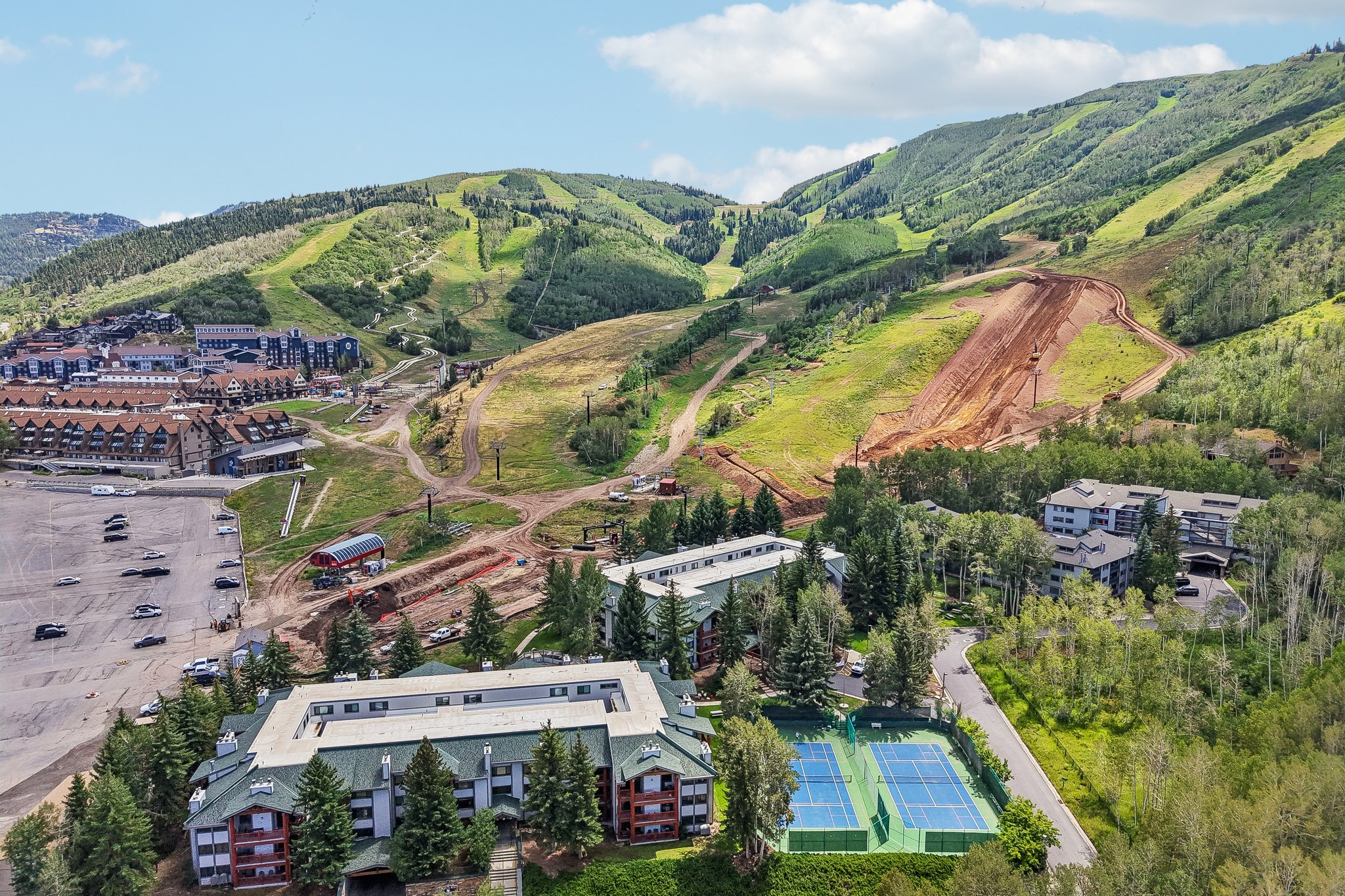 Expansive mountain setting featuring buildings with pitched roofs, surrounded by dense trees, and a tennis court