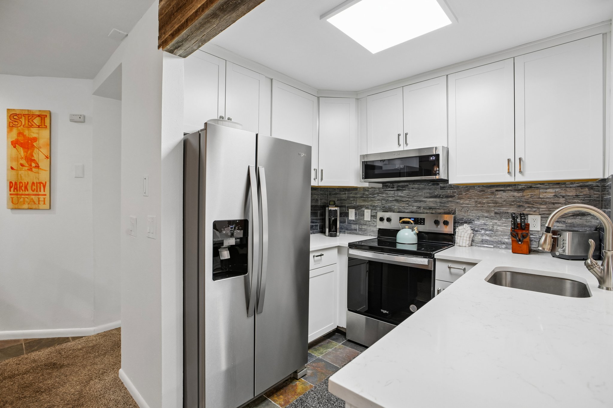 Modern kitchen featuring white shaker cabinetry, stainless steel appliances, a multi-tone tile backsplash, light-toned countertops, and an undermount sink with a gooseneck faucet