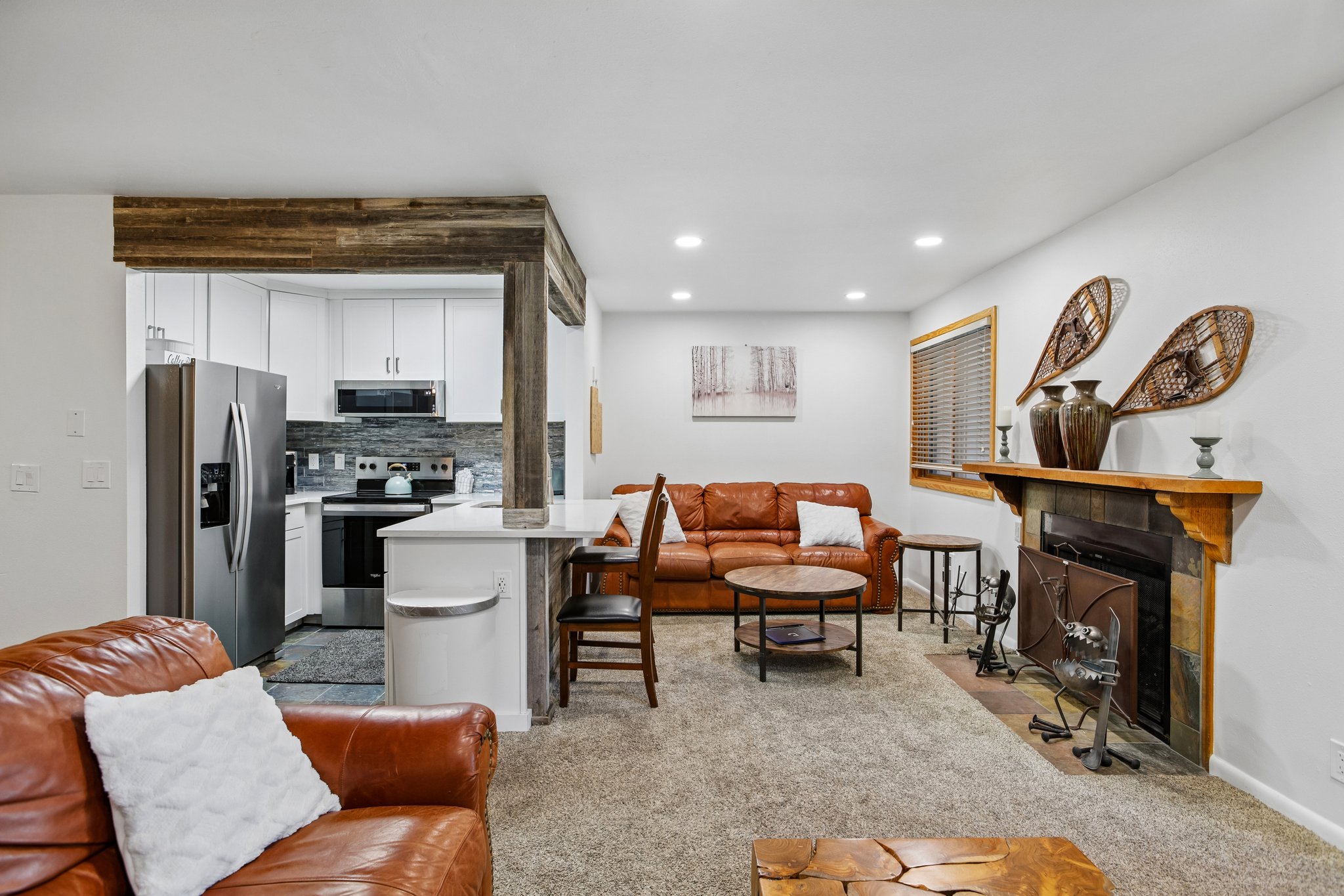 Open-concept layout featuring a kitchen with white cabinetry and stainless steel appliances, a breakfast bar with white countertops, a living area with a stone-surround fireplace and wood mantel, recessed lighting, and wood-finish ceiling accents