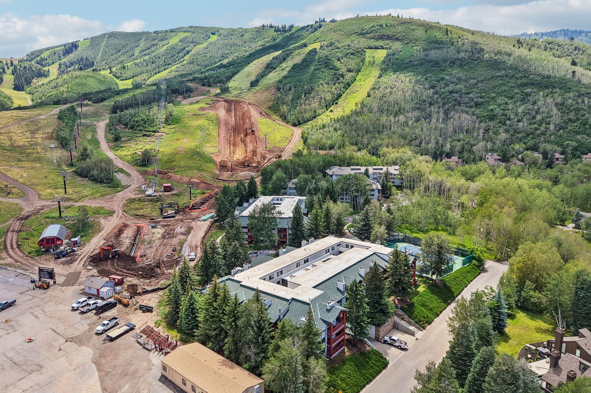 Mountain resort property featuring multiple buildings with green pitched roofs, surrounded by dense evergreen and deciduous trees, with a tennis court visible on the property