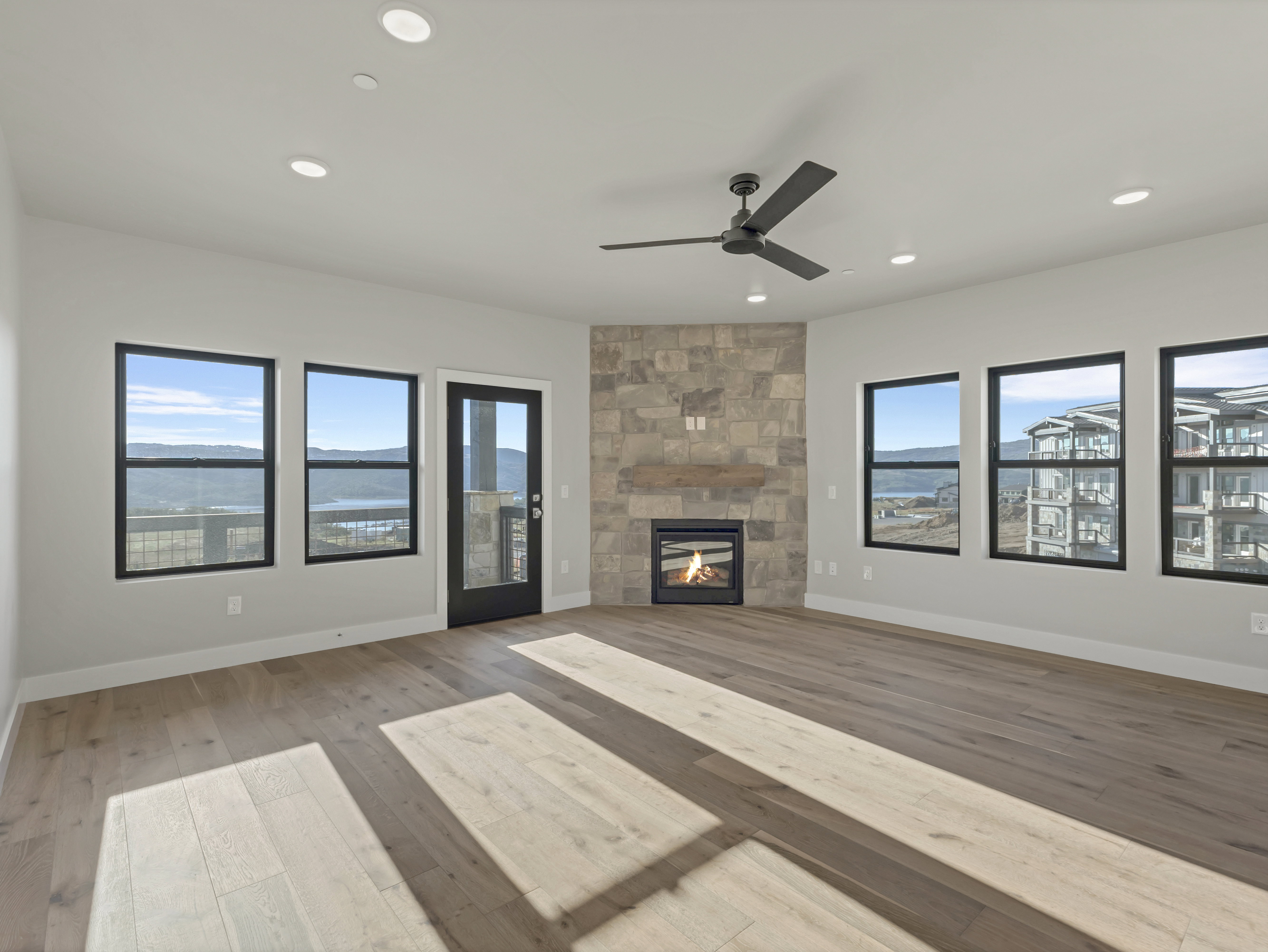 Spacious room featuring wood-finish flooring, a stone-clad fireplace, and multiple windows with black framing