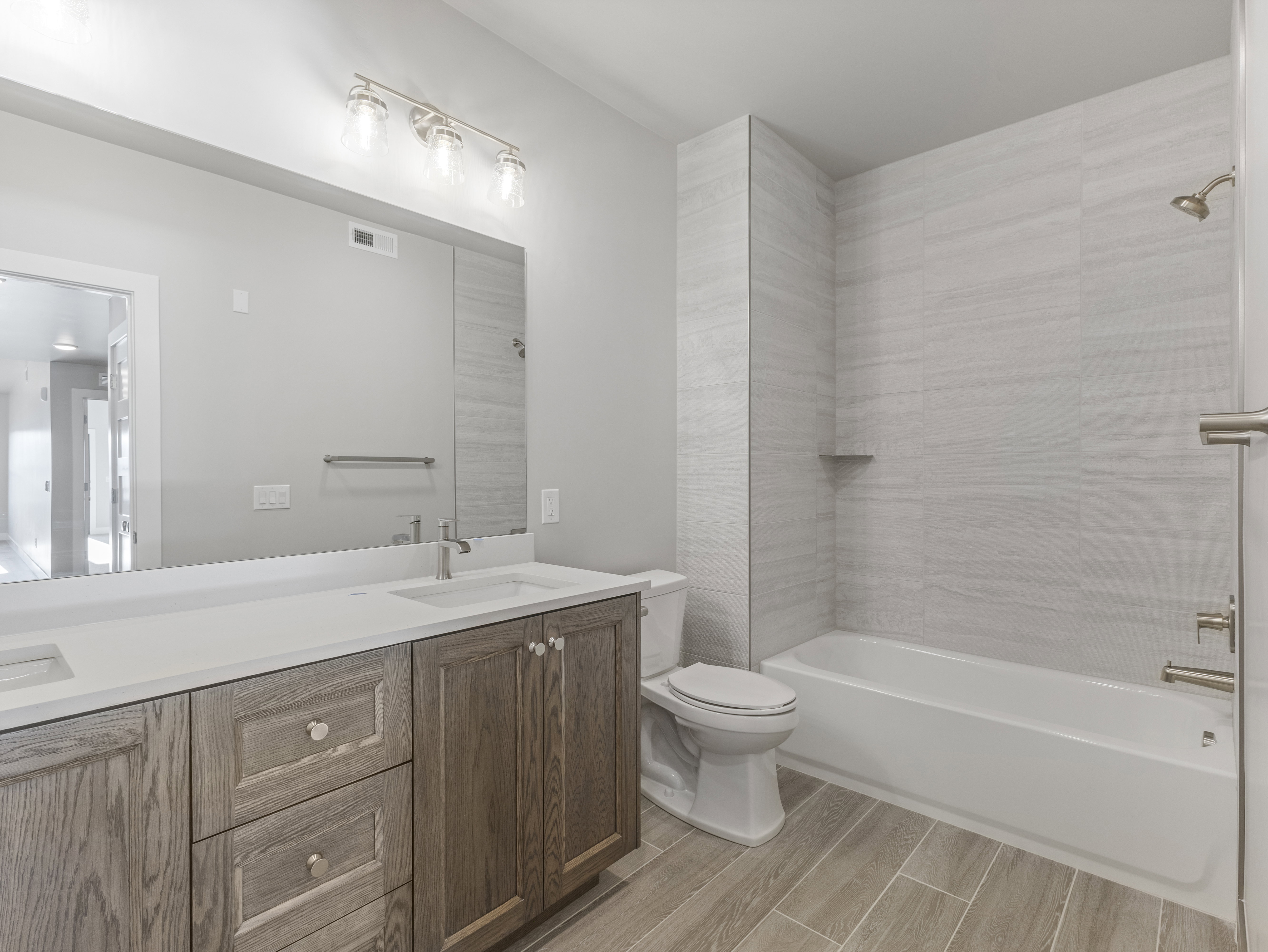 Bathroom featuring a double vanity with white countertops and wood-finish cabinetry, a large wall-mounted mirror, and a full bathtub with tiled surround