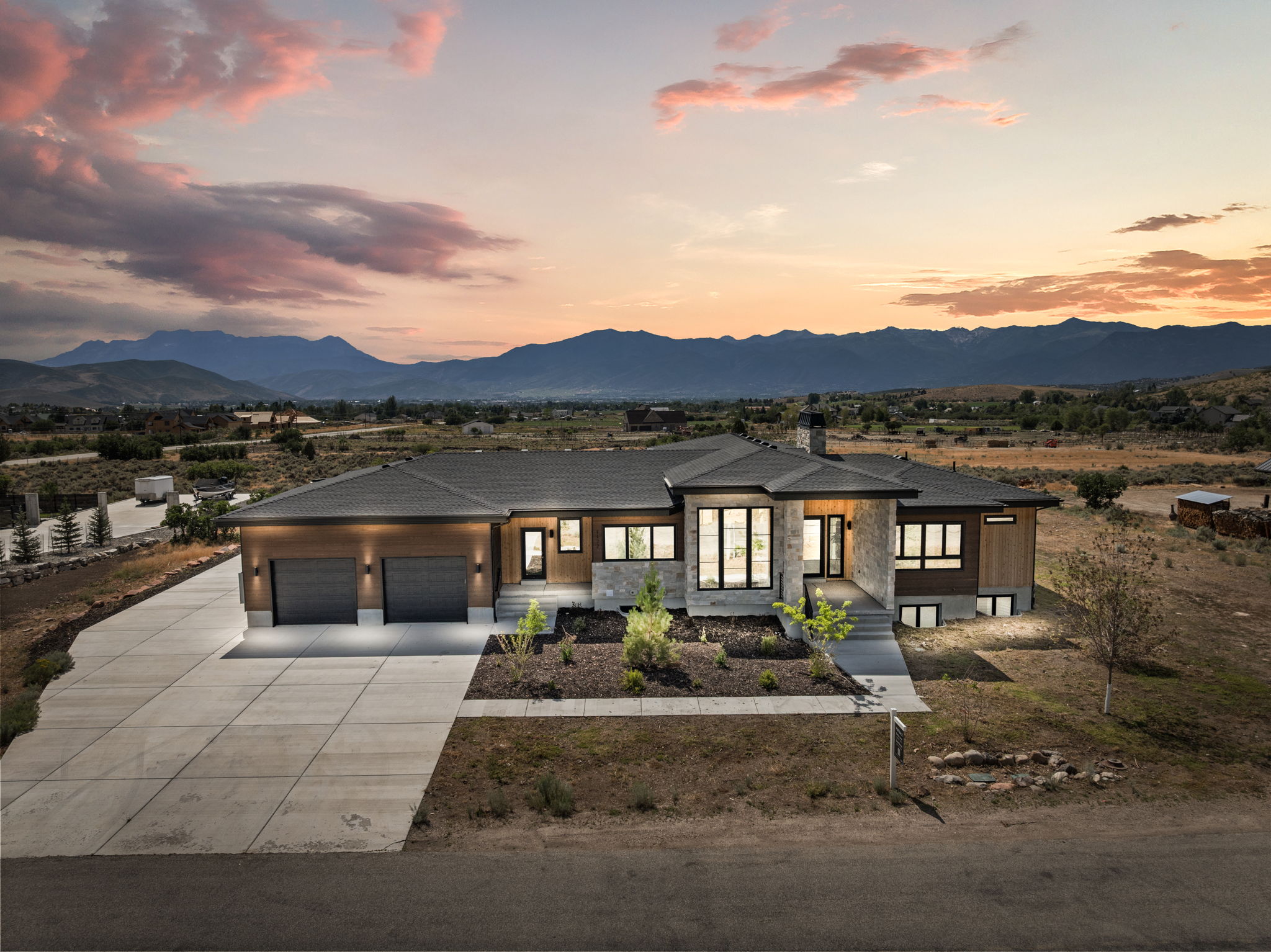 Modern architectural design featuring extensive window placements, a multi-car concrete driveway, a two-car garage, and a stone and wood-paneled exterior