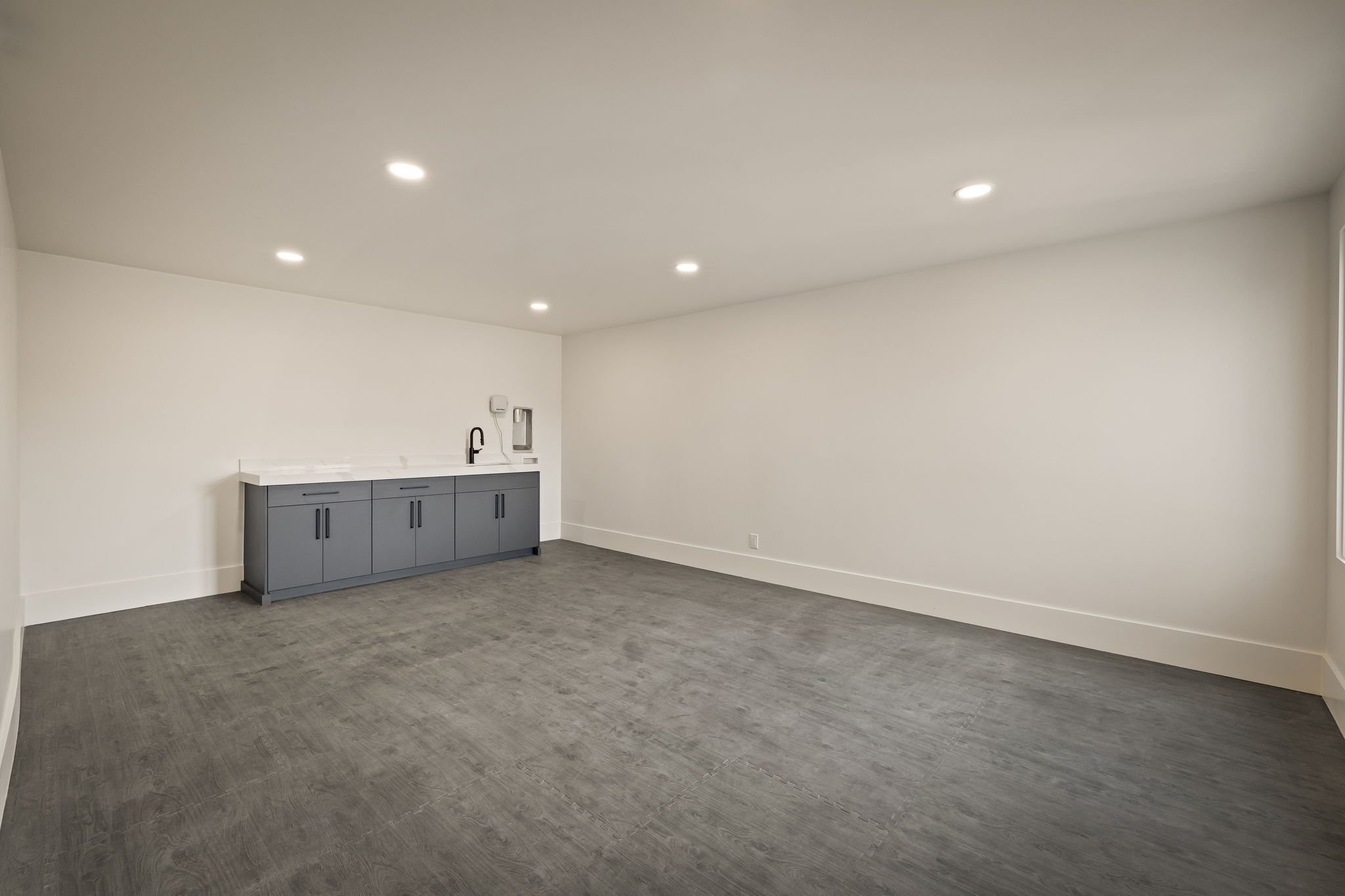 Spacious room featuring dark wood-finish flooring, recessed lighting, and a built-in wet bar with gray cabinetry, a light-toned countertop, and a black faucet
