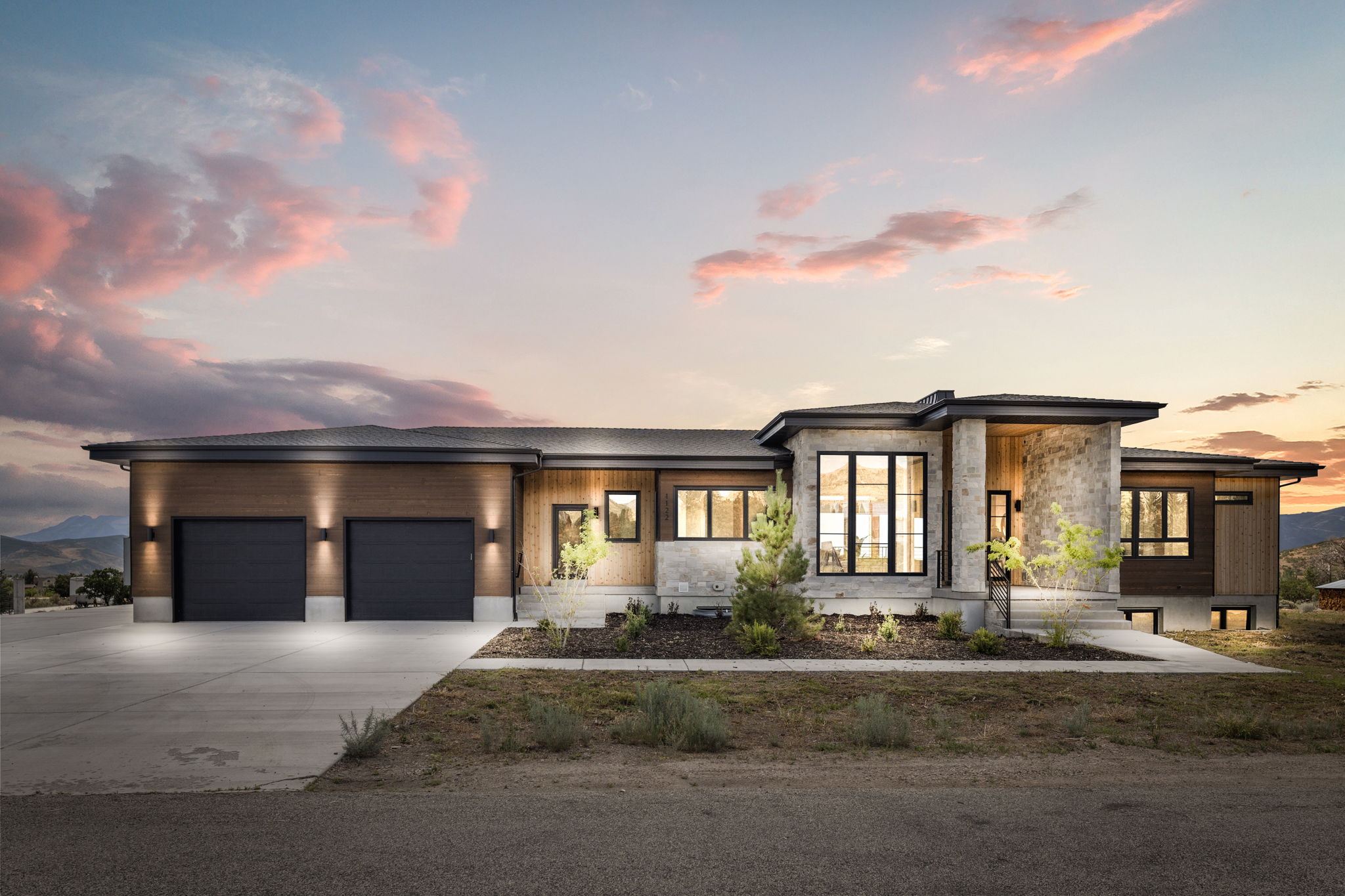 Contemporary exterior featuring stone and wood-look siding, a prominent two-car garage, large black-framed windows, and integrated exterior lighting