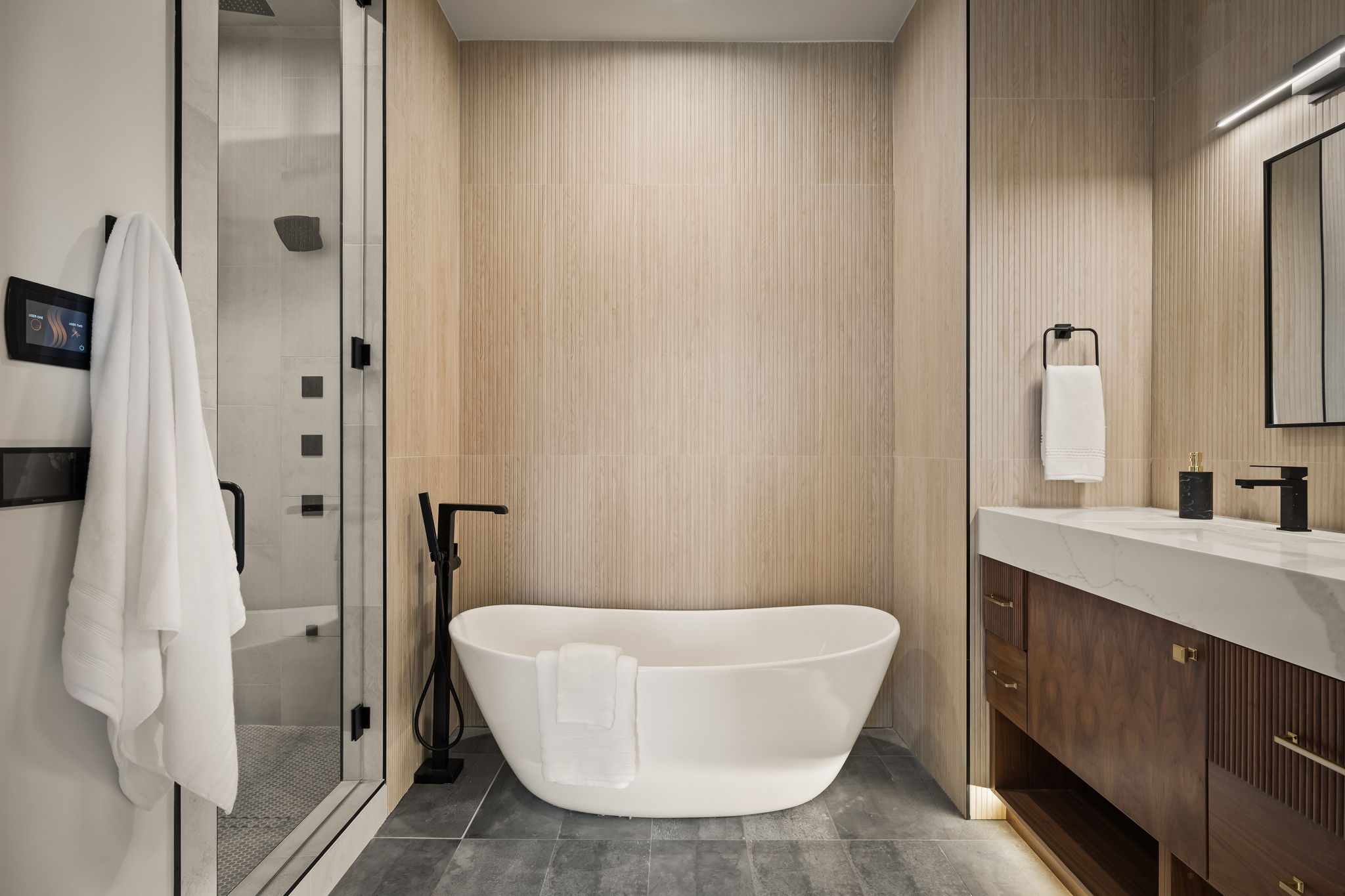 Contemporary bathroom featuring a freestanding soaking tub, a frameless glass-enclosed shower, a wood-finish vanity with a stone countertop, black fixtures, and light-toned wall paneling