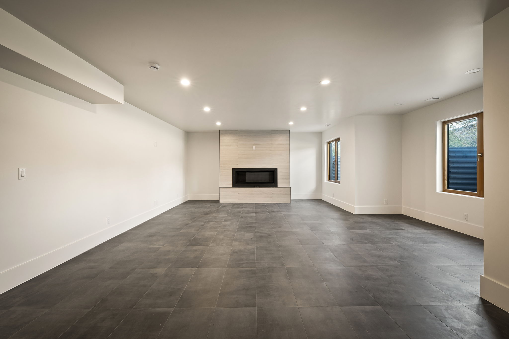 Spacious room featuring dark tile flooring, a modern fireplace with light-toned stone surround, recessed lighting, and multiple windows
