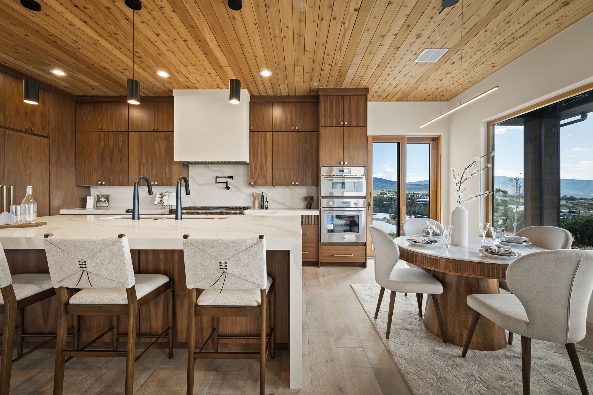 Gourmet kitchen featuring a wood-plank ceiling, integrated wood cabinetry, a large center island with a stone countertop, built-in double ovens, and a dining area with expansive windows