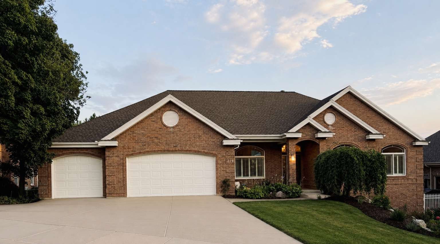 Brick exterior featuring a multi-car garage, pitched roof, arched entry, and mature landscaping