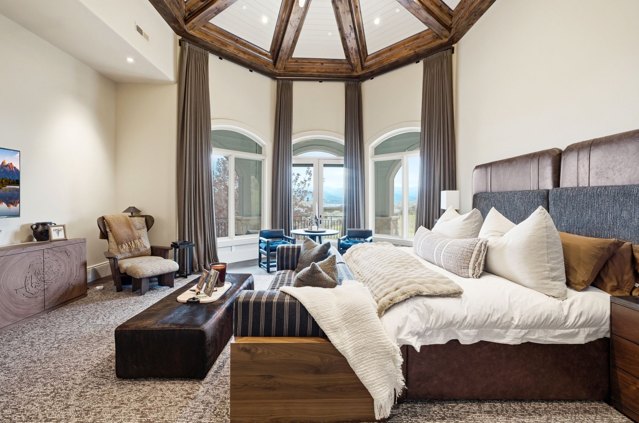 Spacious room featuring vaulted ceilings with exposed wood beams, a large bed with a multi-textured headboard, and a sitting area with multiple windows