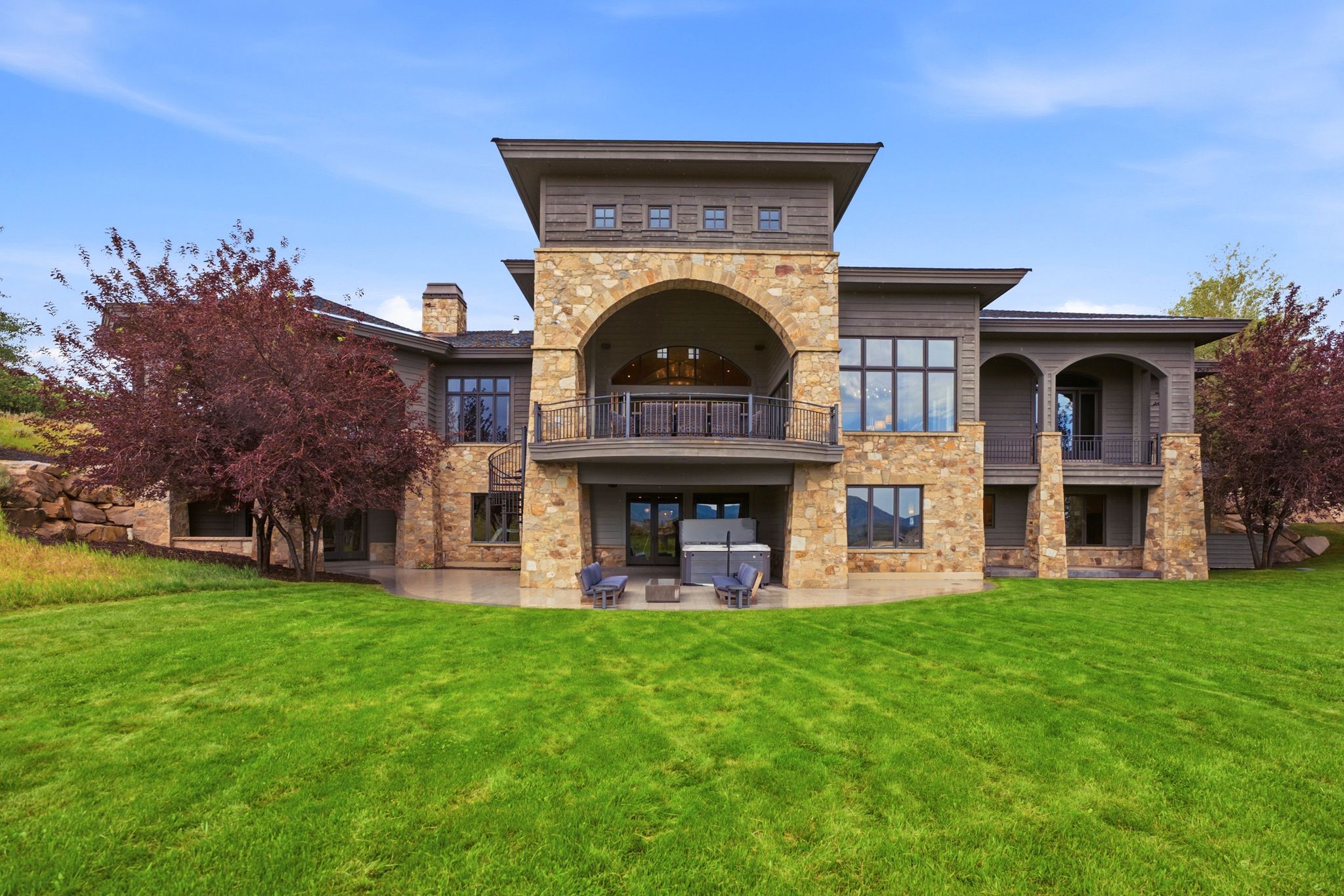 Expansive rear exterior featuring a stone and wood facade, multiple balconies, a lower-level patio, and a vast lawn
