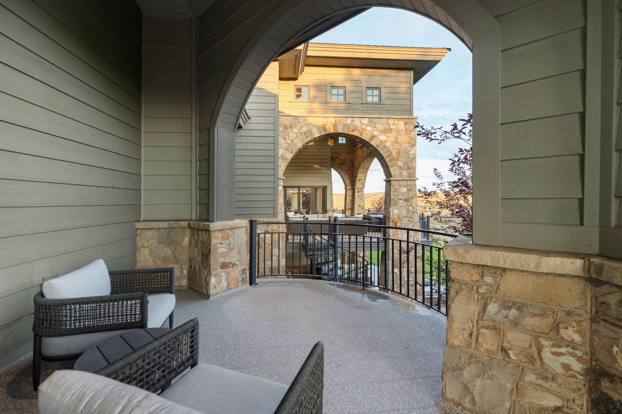 Covered outdoor patio featuring a textured concrete surface, arched architectural details, stone-clad pillars, and a black wrought-iron railing