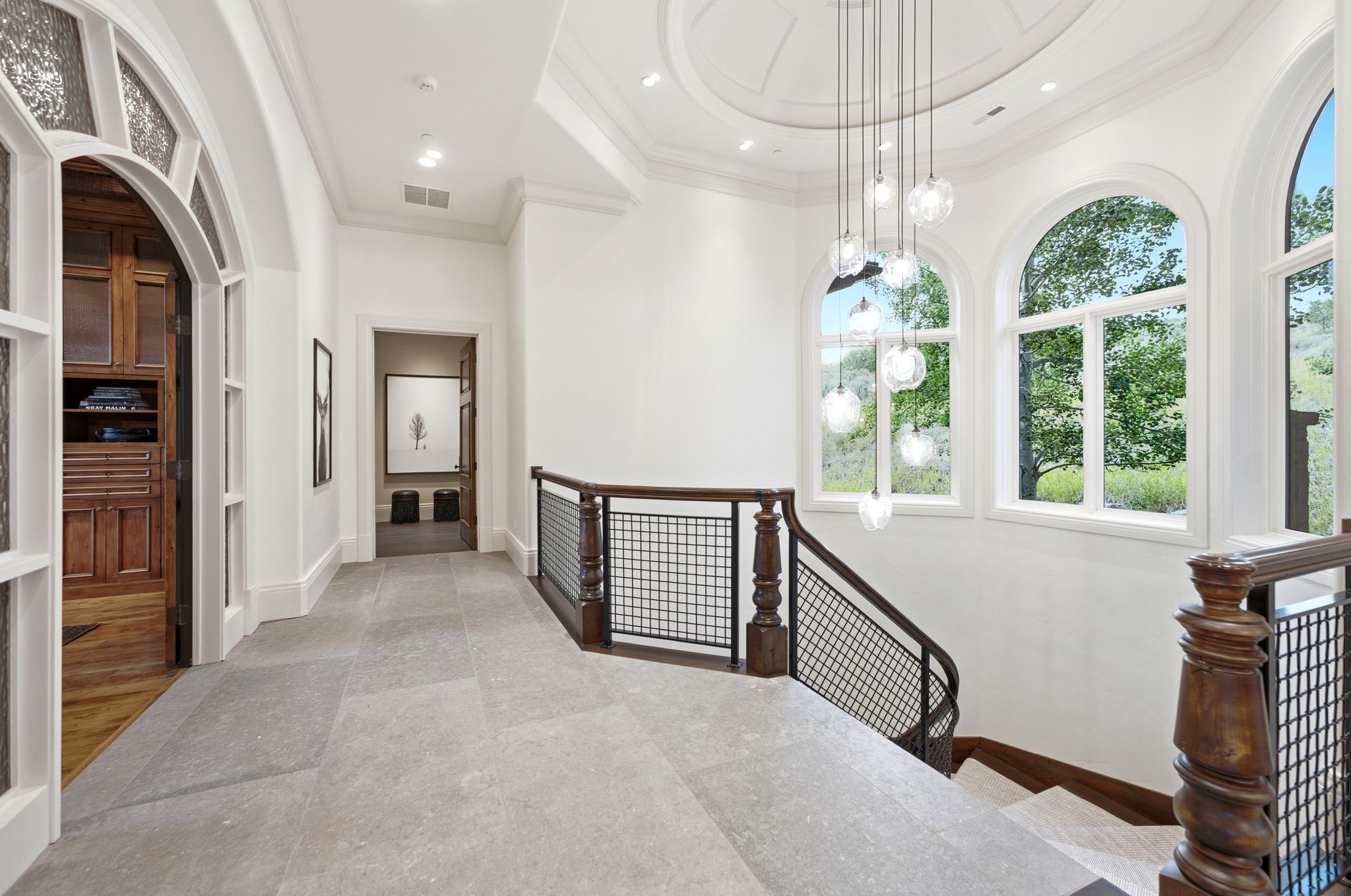 Gallery-style hallway featuring light-toned flooring, a decorative chandelier with multiple glass globes, and arched windows providing outdoor views