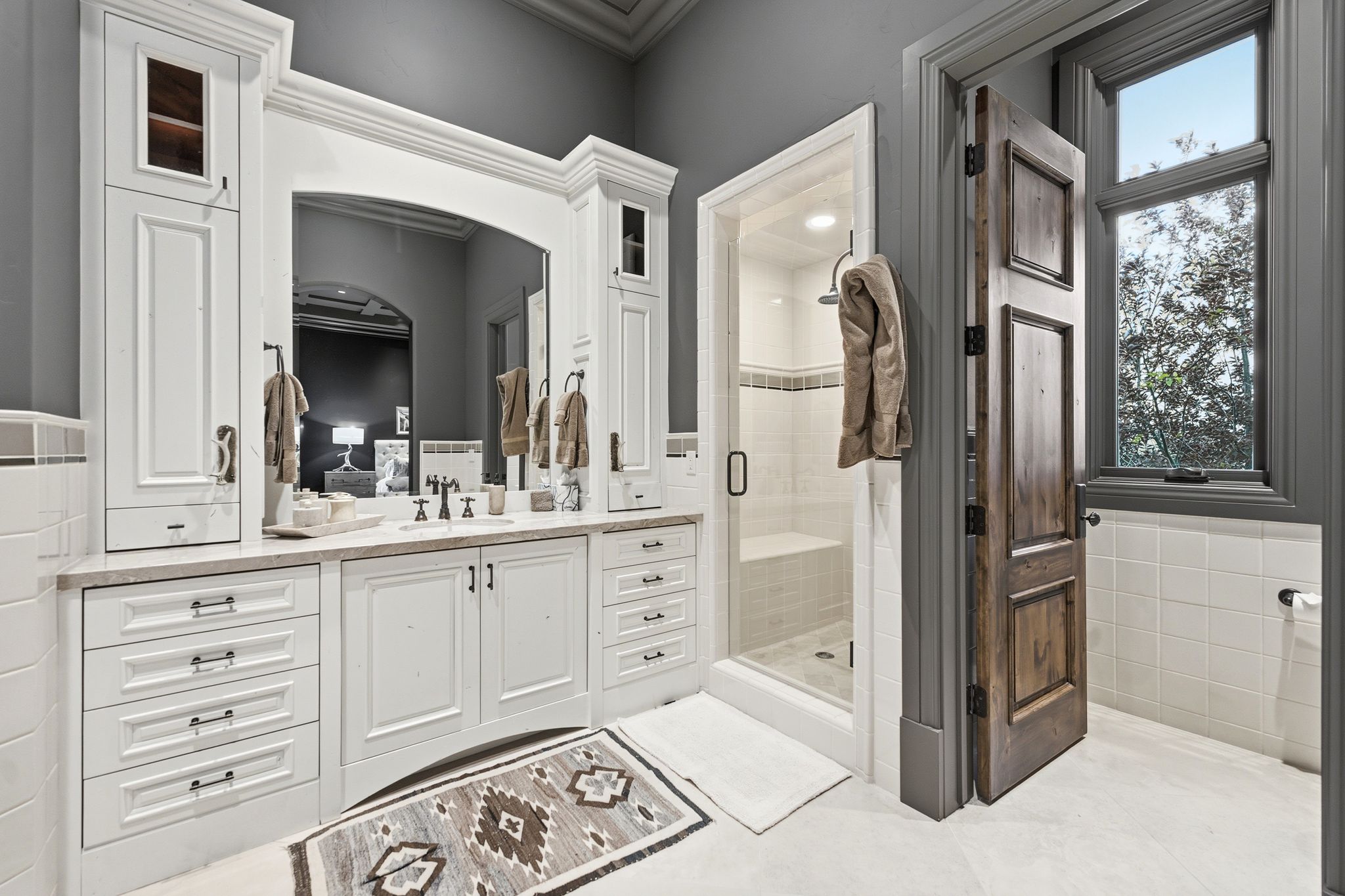 Bathroom featuring a dual vanity with light-toned countertops, white shaker-style cabinetry, and an integrated glass-enclosed shower with a bench