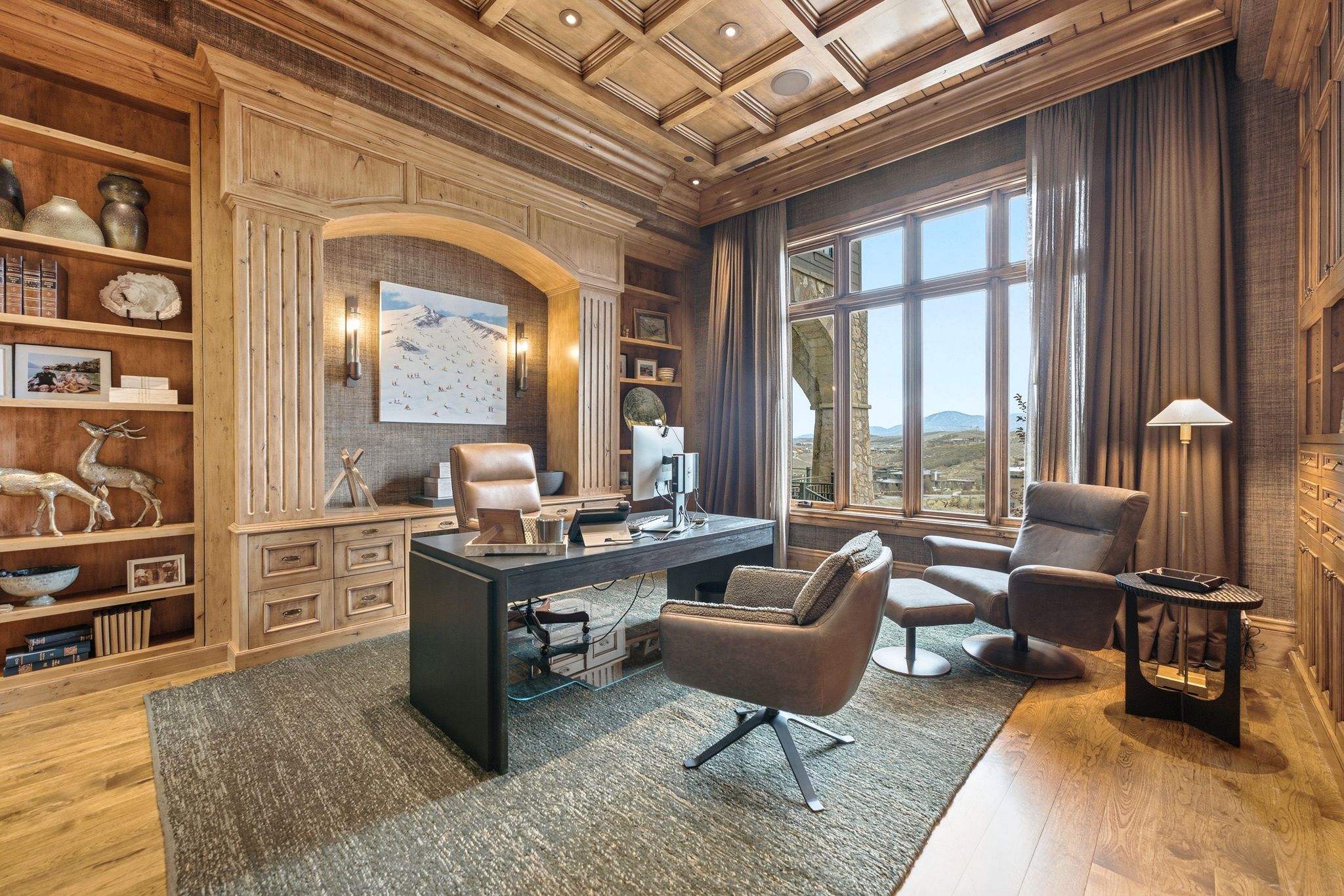 Wood-paneled study featuring a coffered ceiling, built-in shelving, architectural archway with sconces, and a large multi-pane window overlooking a mountainous landscape