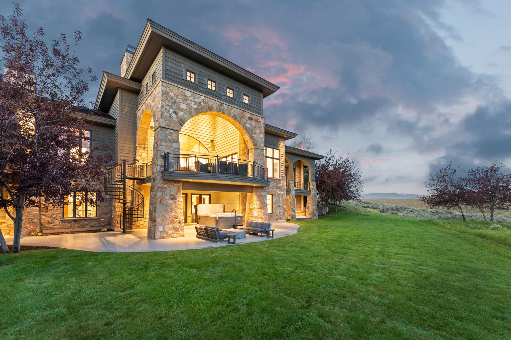Stone and siding exterior with arched balcony, lower patio, spiral staircase, and expansive lawn